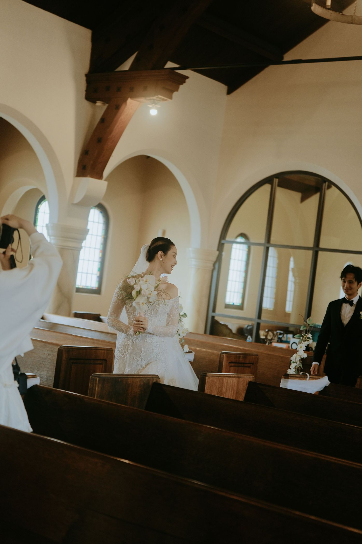 Elegant bride in lace gown smiles holding bouquet inside a charming white arched church interior.