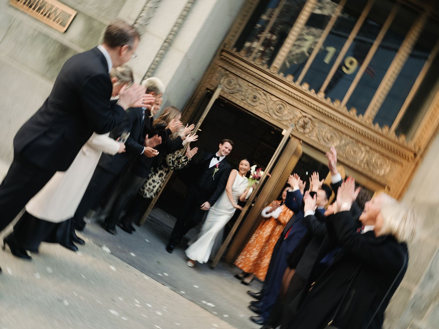 Blurred motion photo of wedding guests cheering as couple exits grand ceremonial building entrance.