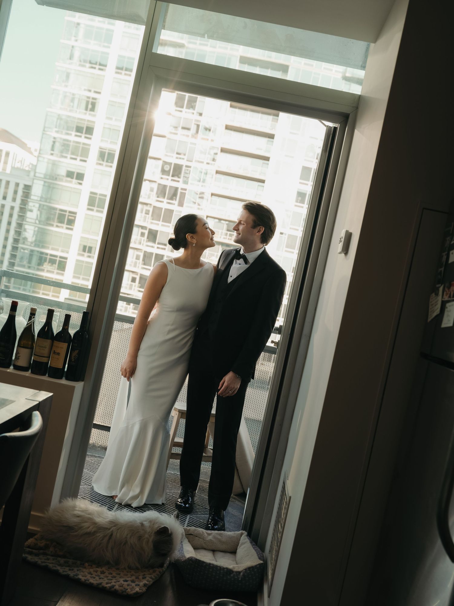 Newlyweds standing in a sunlit apartment balcony doorway, smiling at each other in wedding attire.