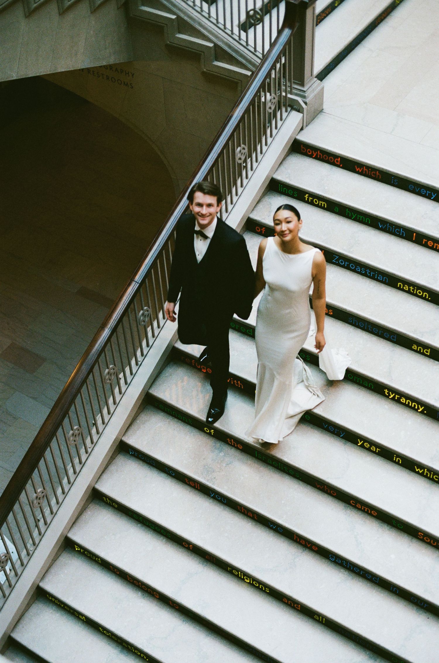Newlyweds descend ornate staircase with gold-lettered steps, bride in white gown and groom in tuxedo.
