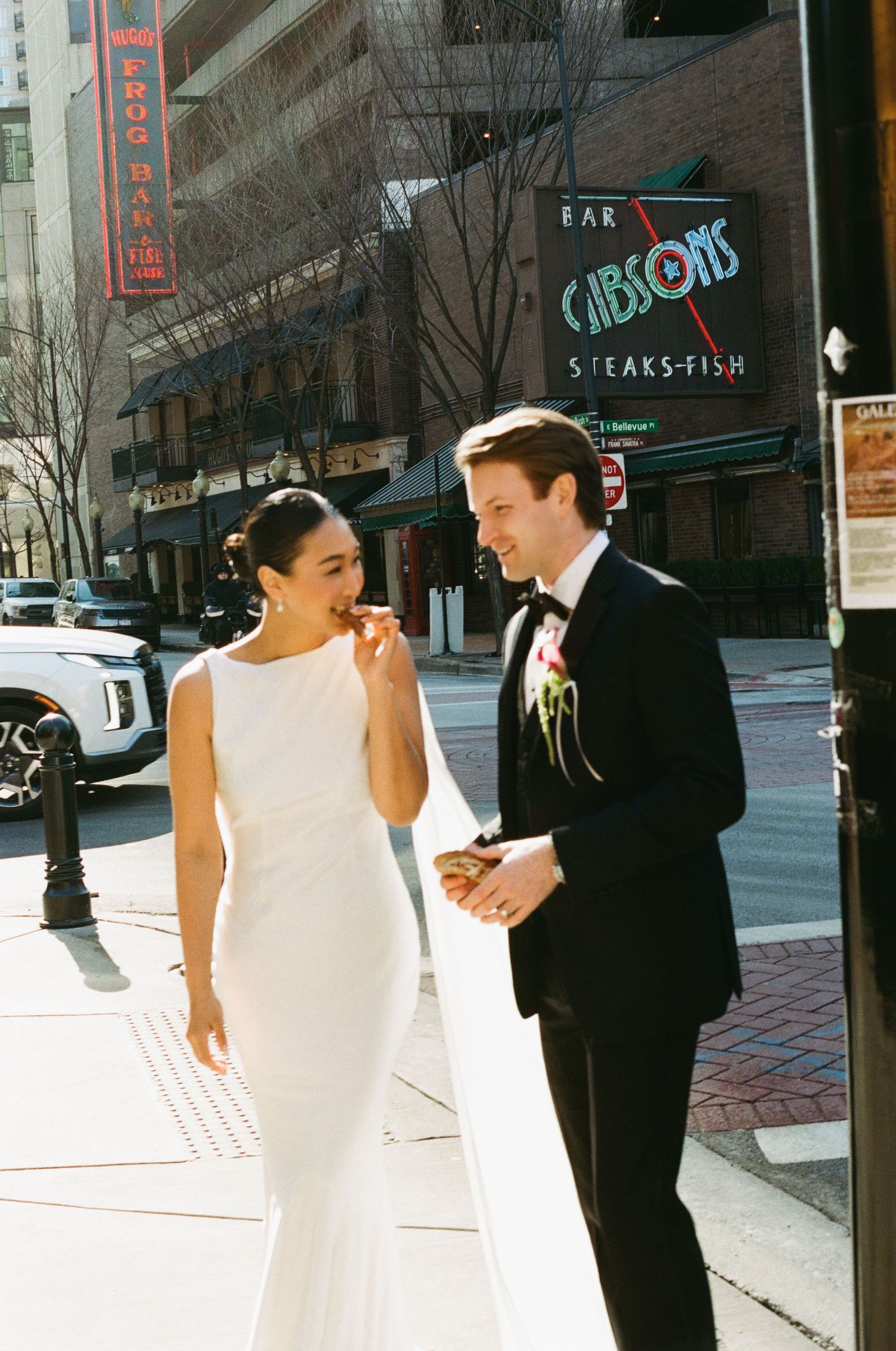 Smiling bride and groom in wedding attire eating snacks outside Gibsons Bar and Steakhouse, Chicago.