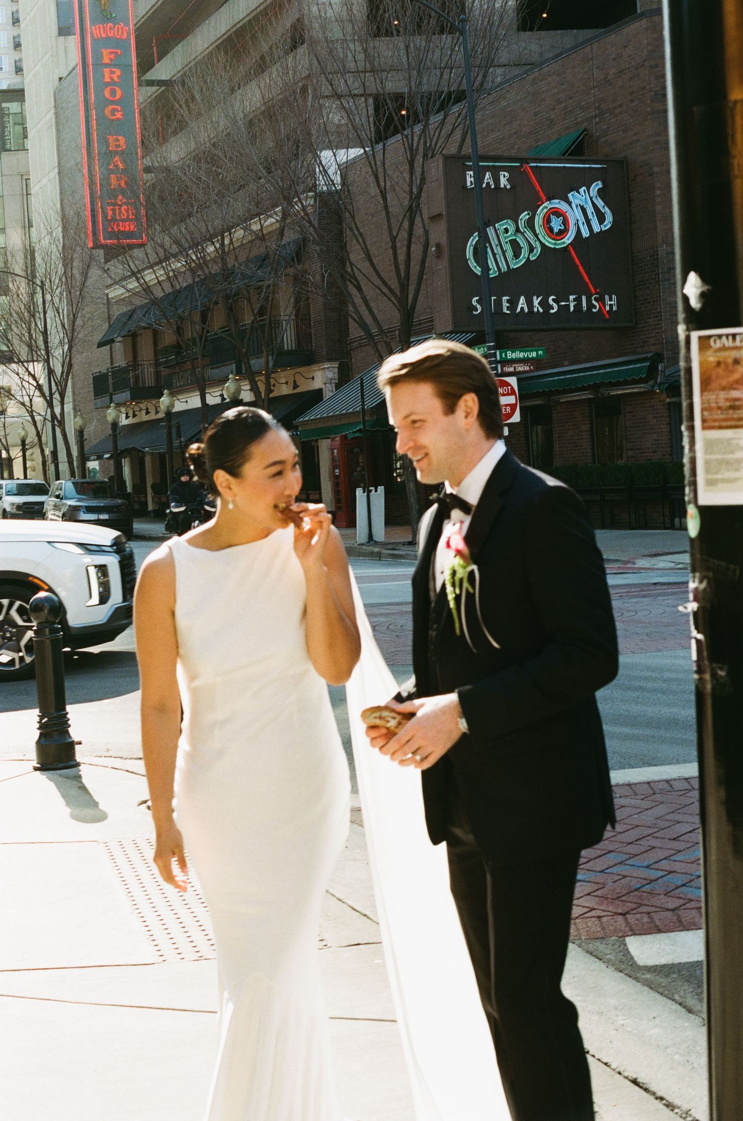 Smiling bride and groom in wedding attire enjoy snacks outside Gibsons Bar and Steakhouse in Chicago.