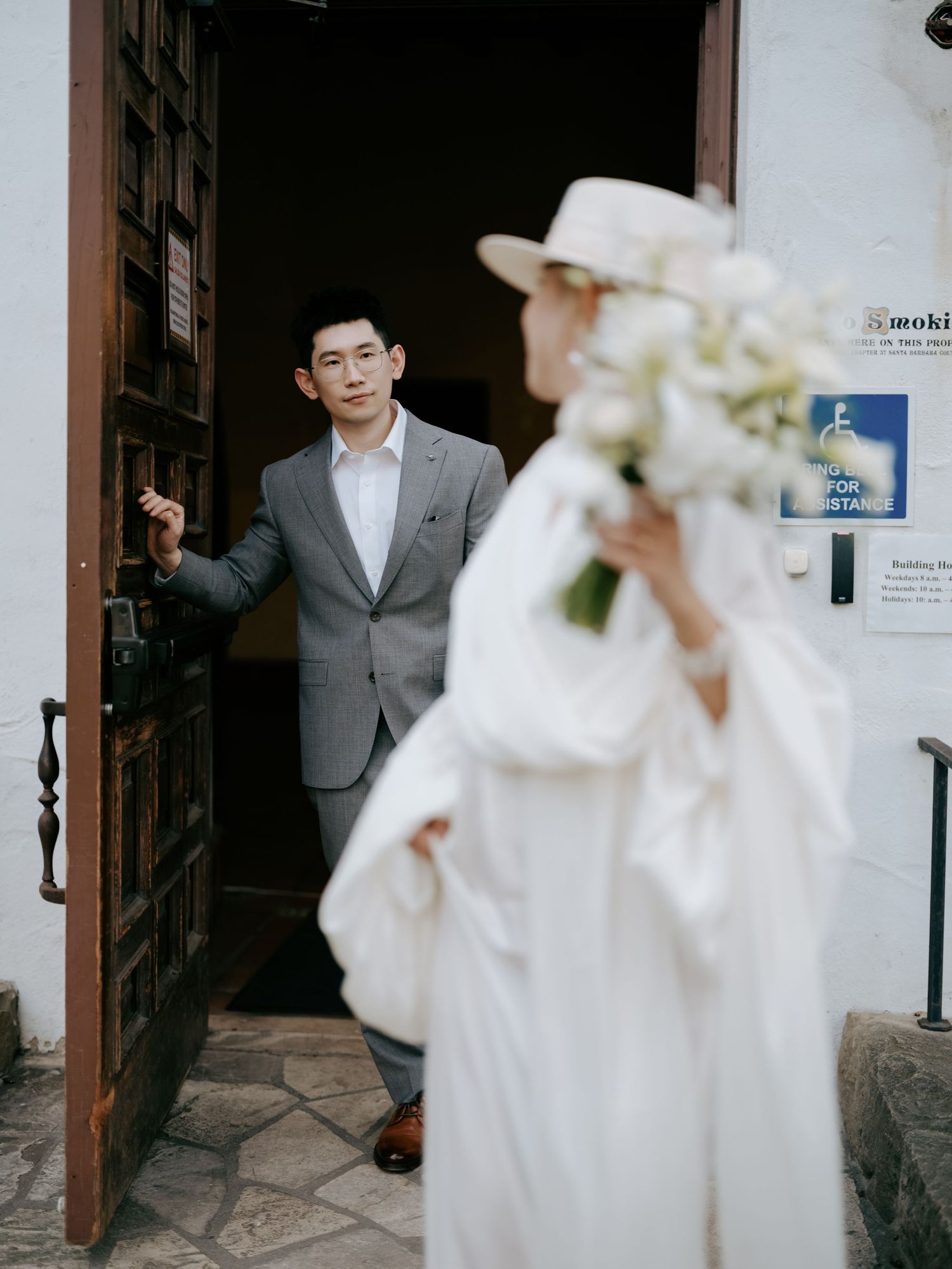 Groom in gray suit holds door open for bride in white cape dress and wide-brim hat.