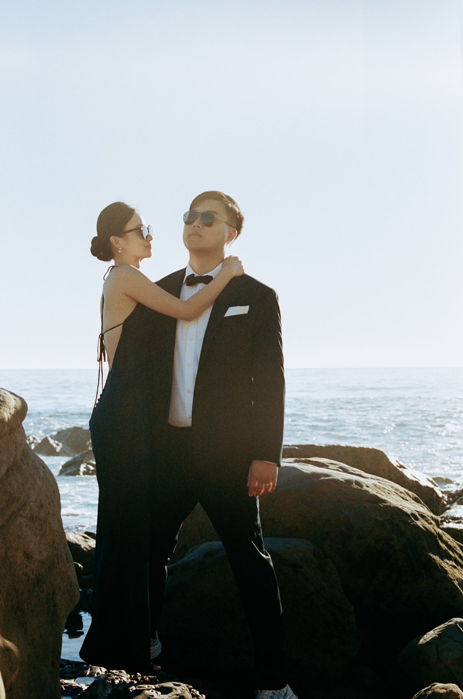 Couple in black formal wear posing on rocky beach, woman adjusting man's bow tie.