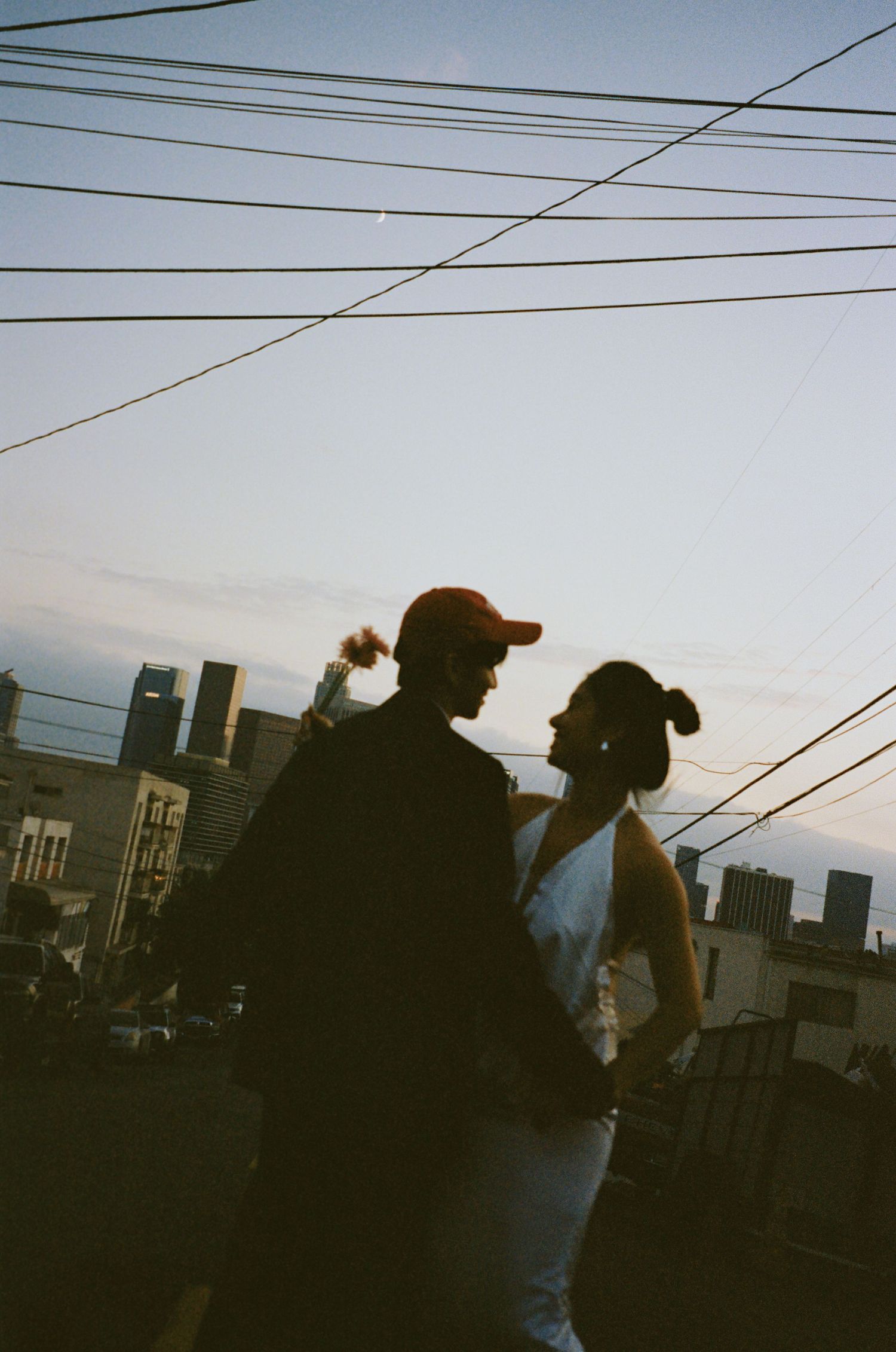 Silhouette of couple standing close together on rooftop at sunset with city skyline and power lines above.