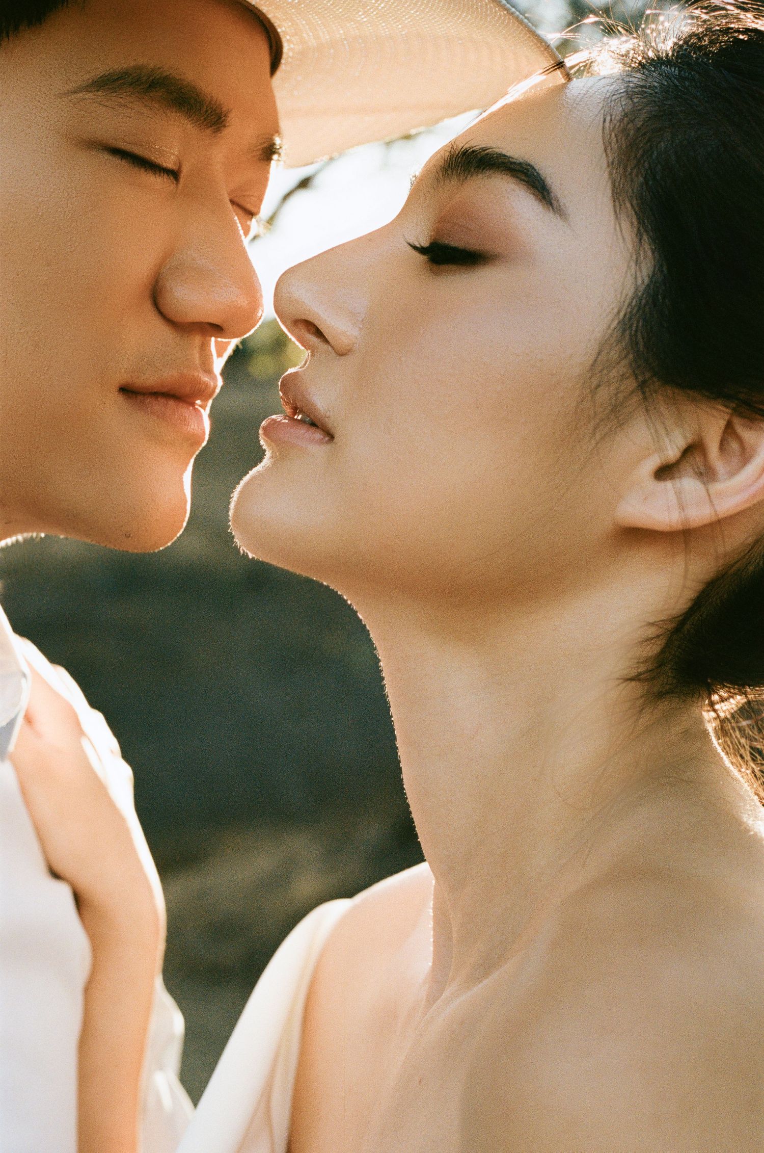 Close-up of an Asian couple almost kissing, bathed in warm golden sunlight outdoors.
