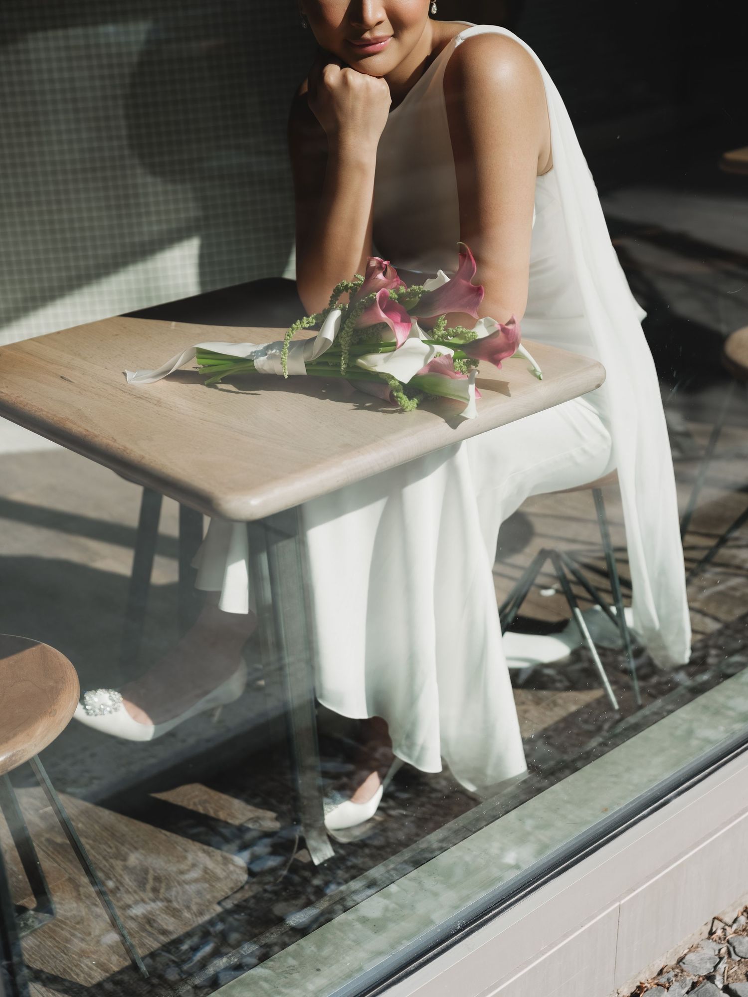 Bride in sleeveless white gown sits at a cafe table with pink flowers, photographed through glass.
