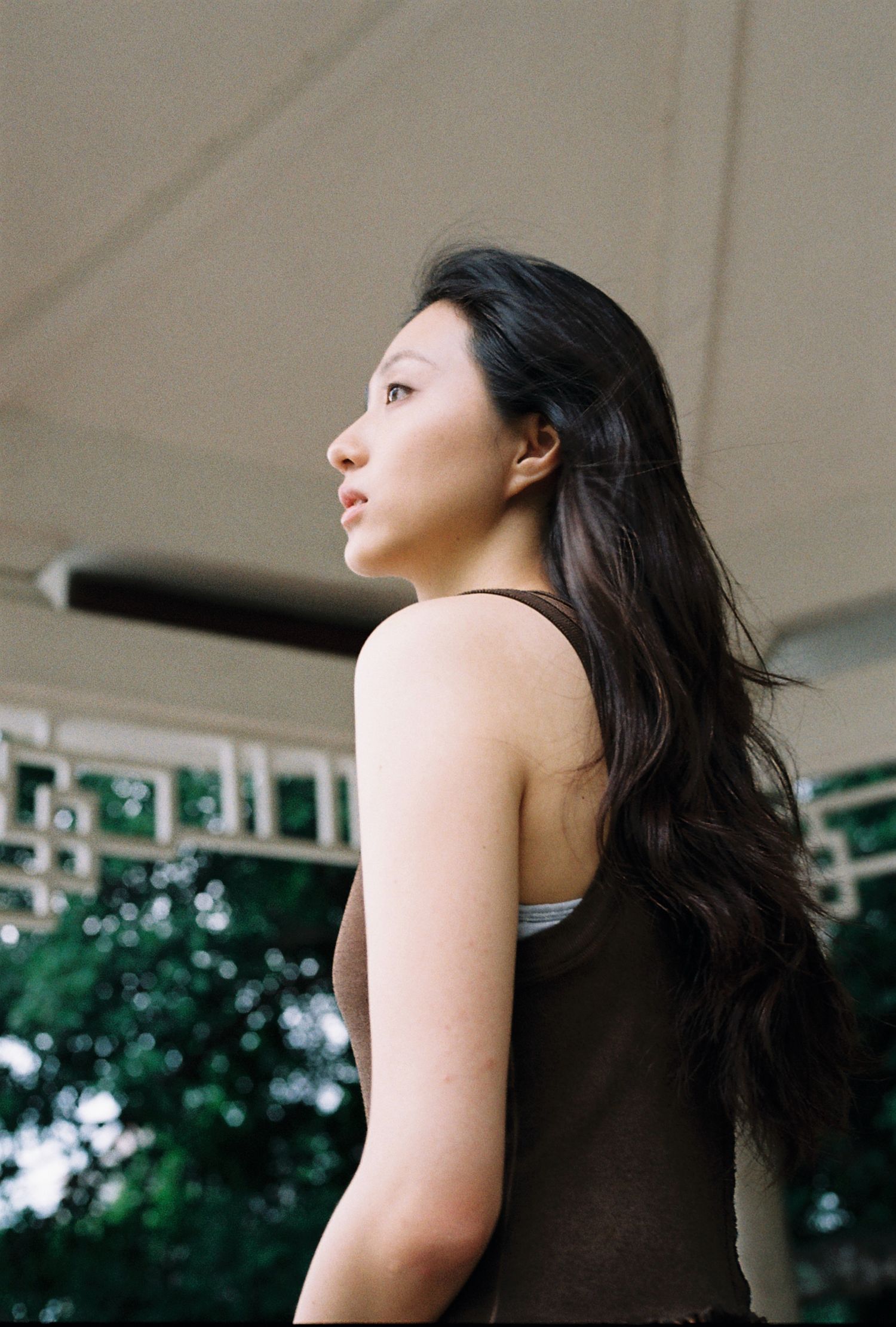 Young Asian woman with long wavy hair looks away in a brown tank top under an outdoor pavilion.