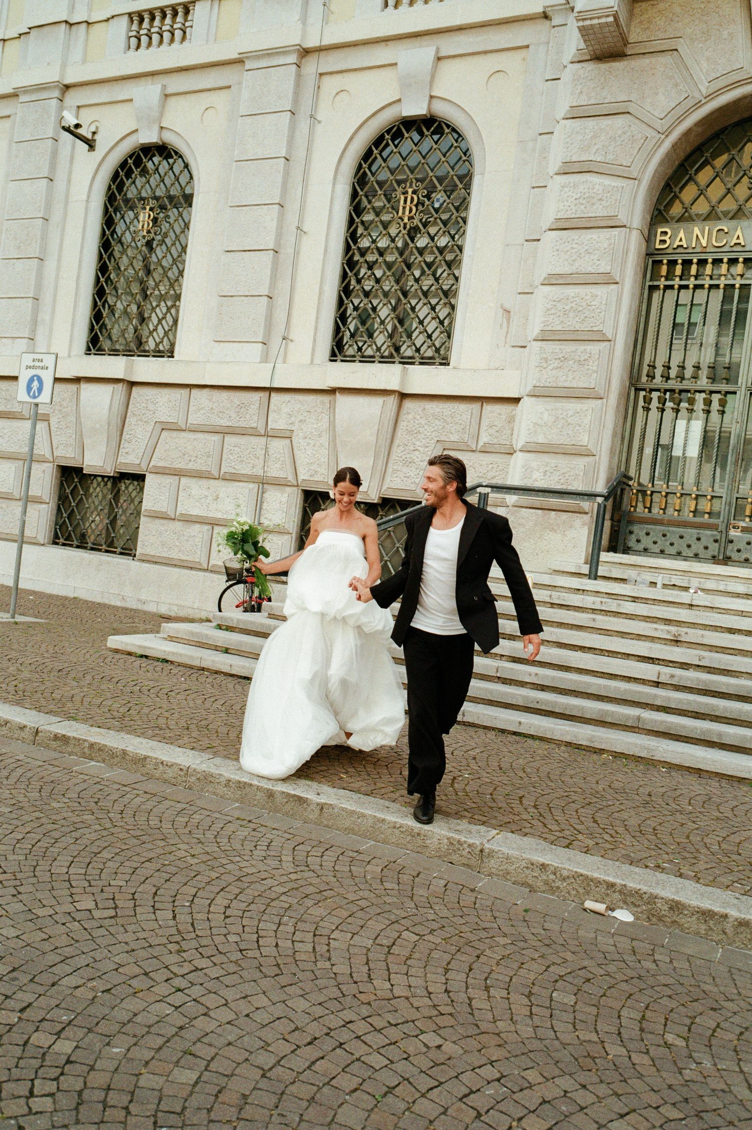 Smiling bride and groom run together on cobblestone street outside a neoclassical Italian building.