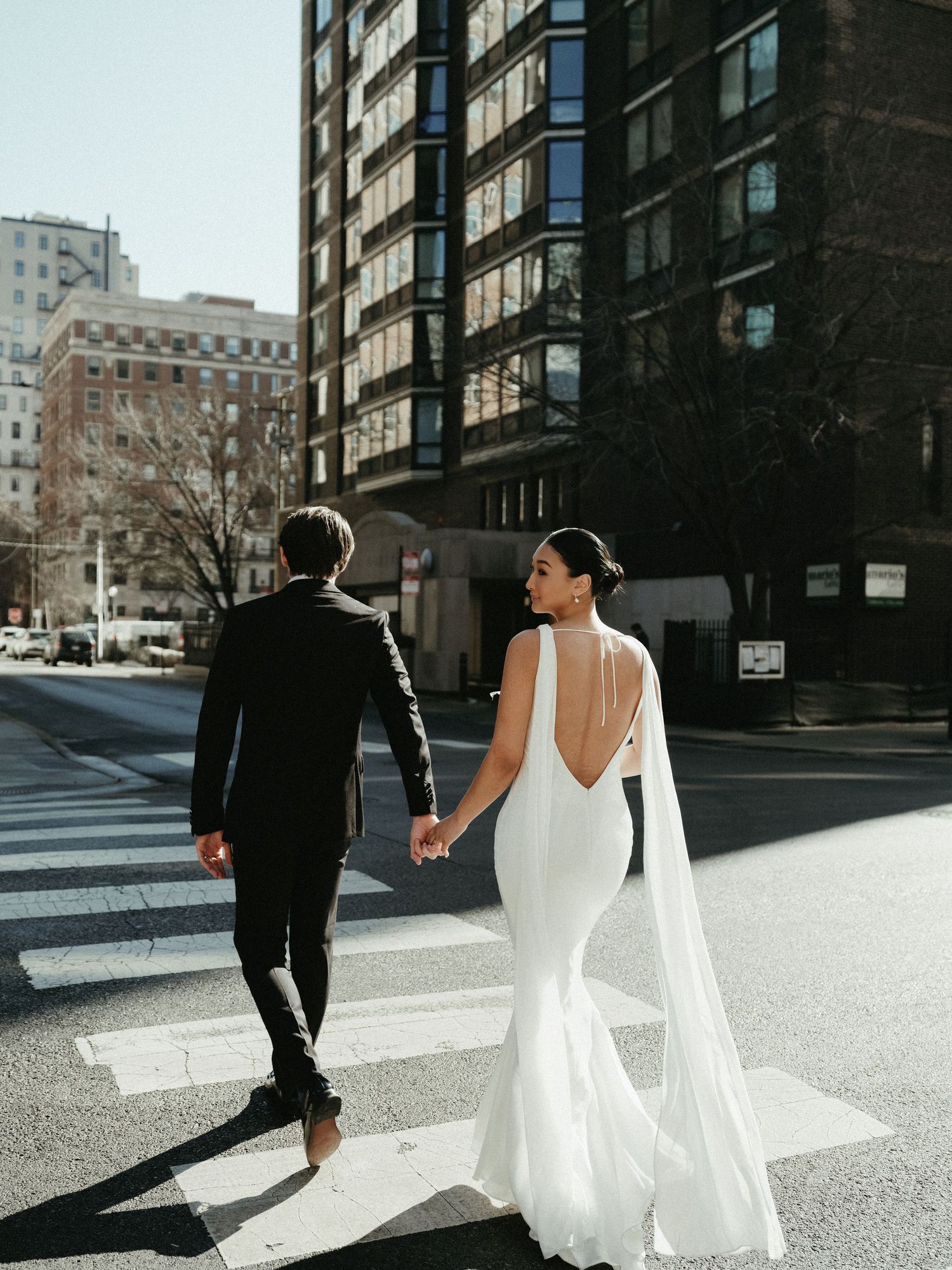 Bride in backless white gown with flowing cape holds grooms hand crossing city street crosswalk together.