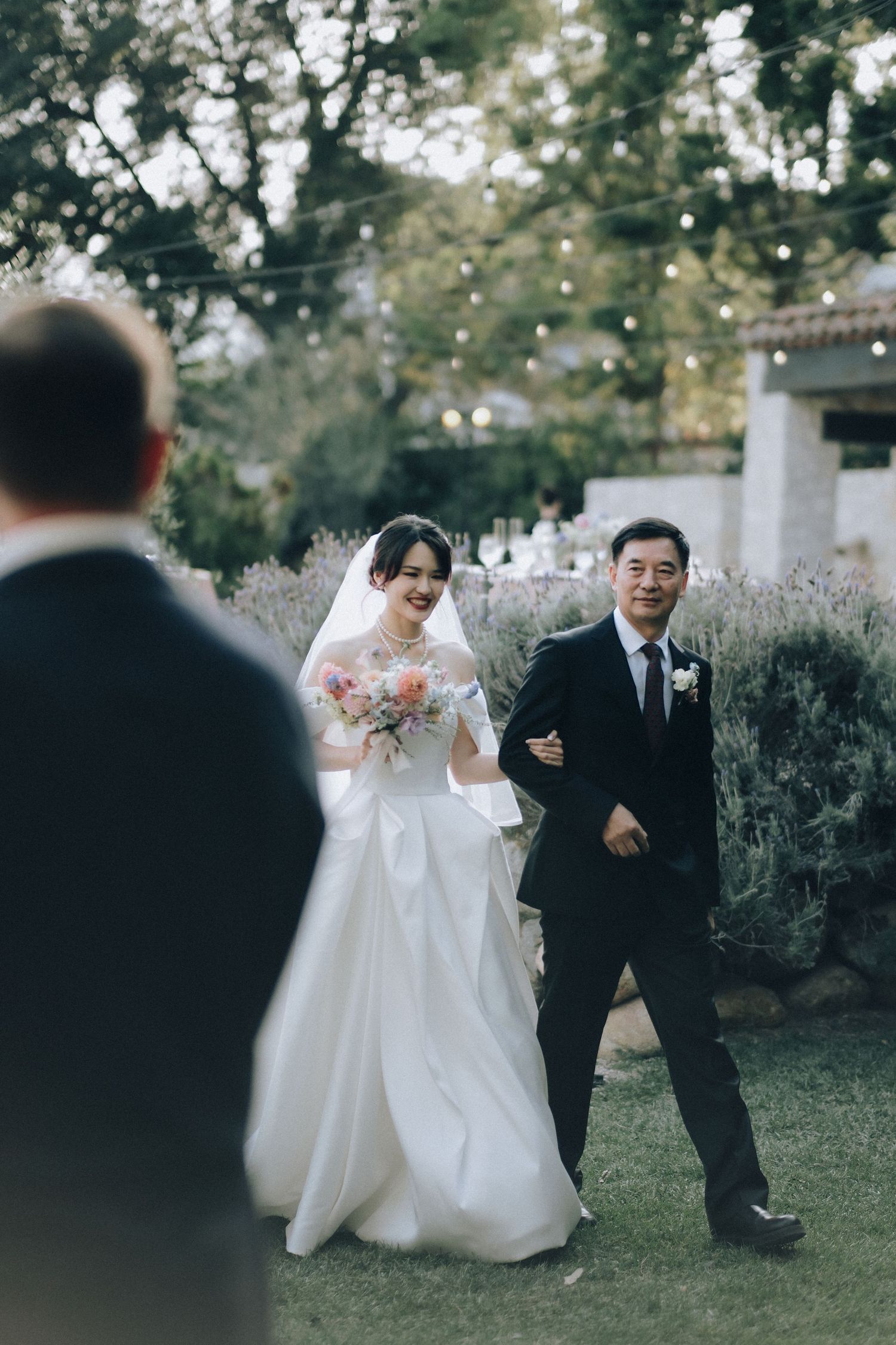 Smiling bride walks down aisle arm-in-arm with father toward groom under string lights outdoors.