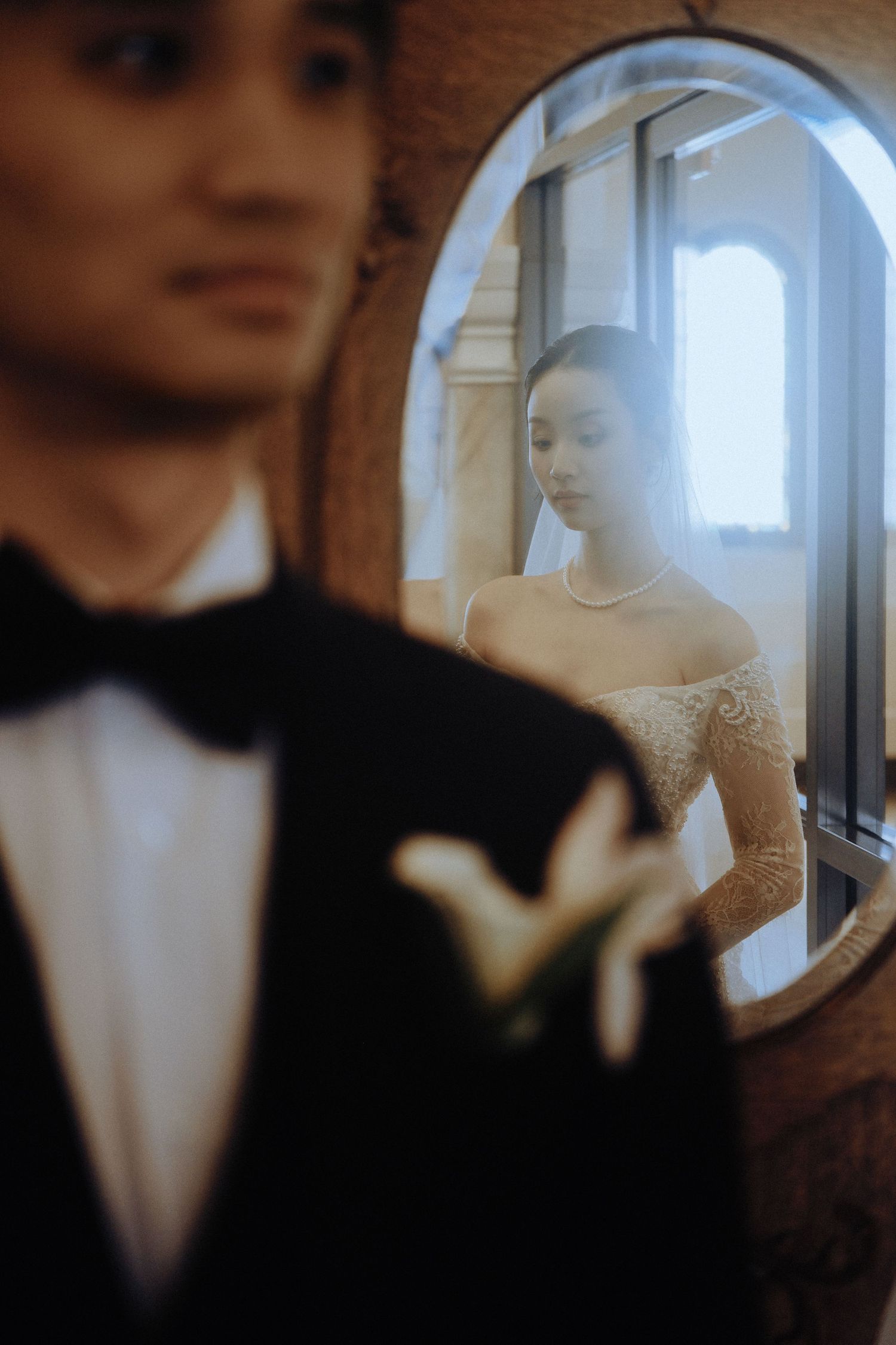 Bride in lace dress reflected in oval mirror as groom stands in foreground.