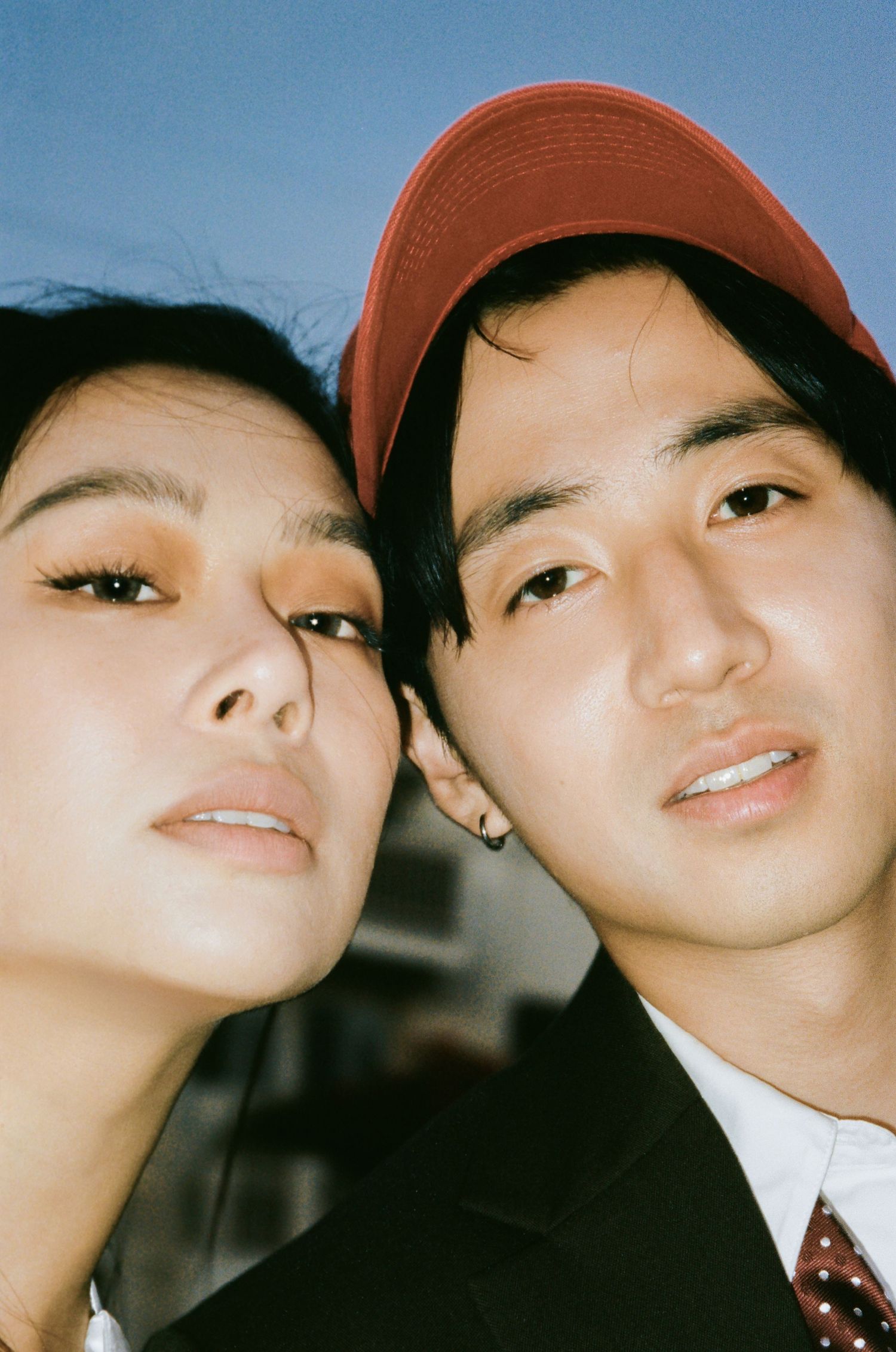 Close-up portrait of Asian couple outdoors, woman with dramatic eye makeup, man wearing red baseball cap.