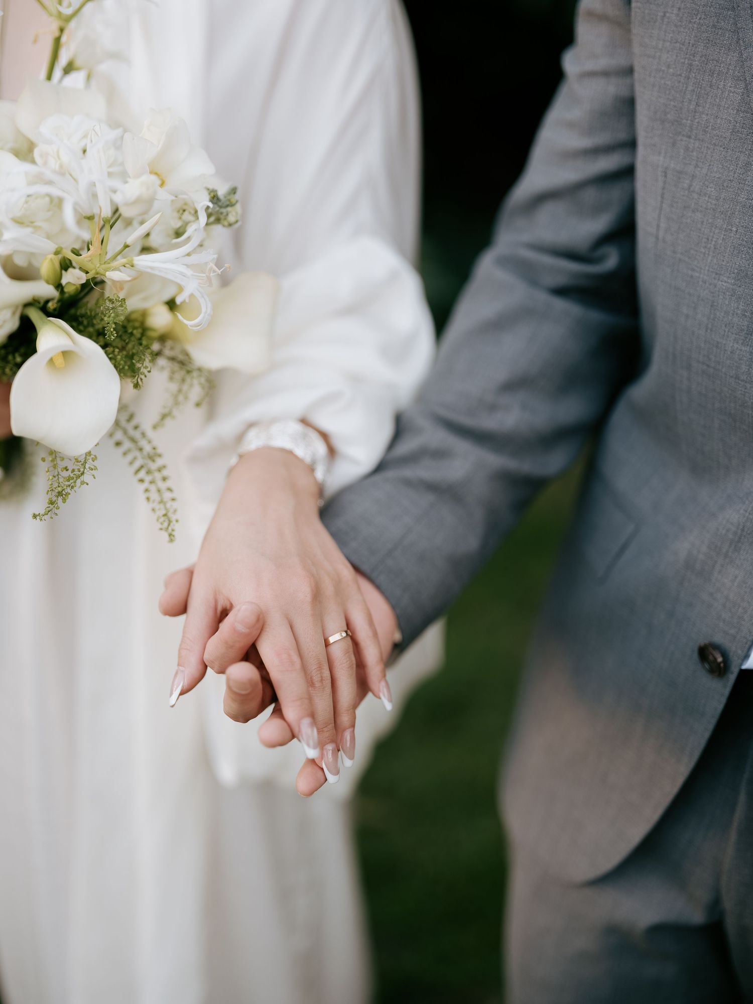 Close-up of newlyweds holding hands with wedding rings, bride holding white floral bouquet.