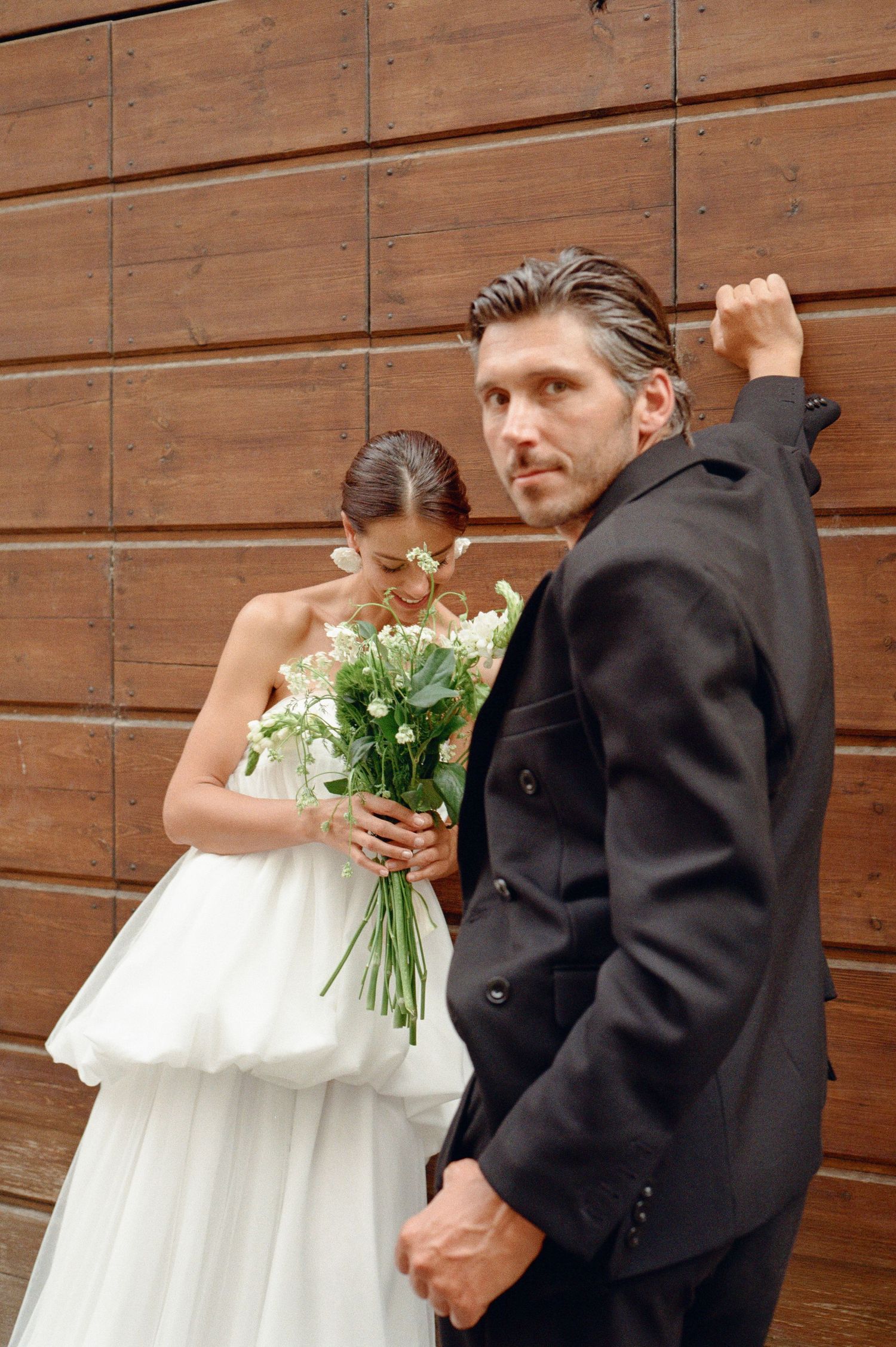 Groom in black suit poses with bride holding white floral bouquet against wooden wall.