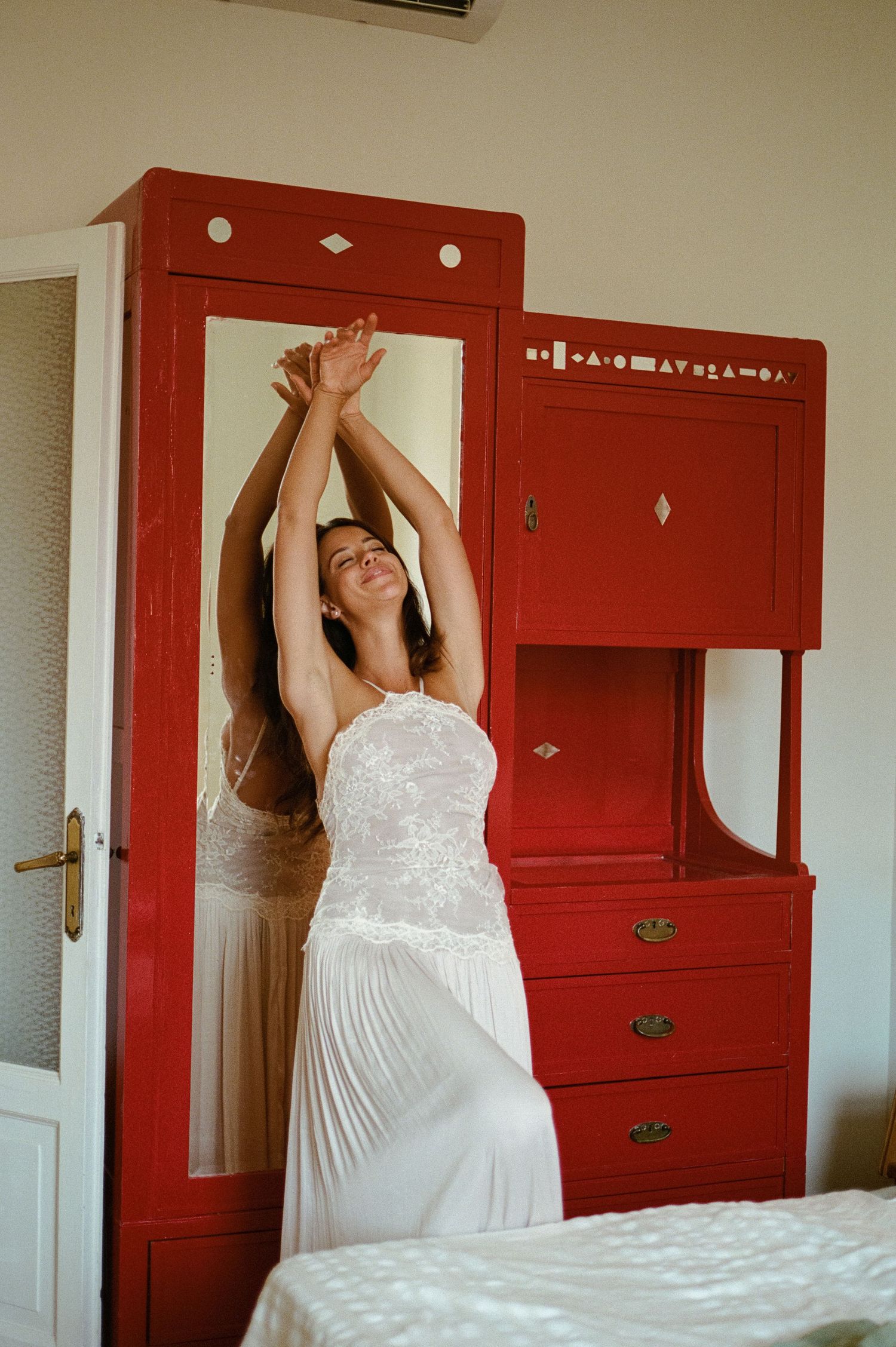 Woman in a white lace dress stretching joyfully in front of a vintage red wardrobe.