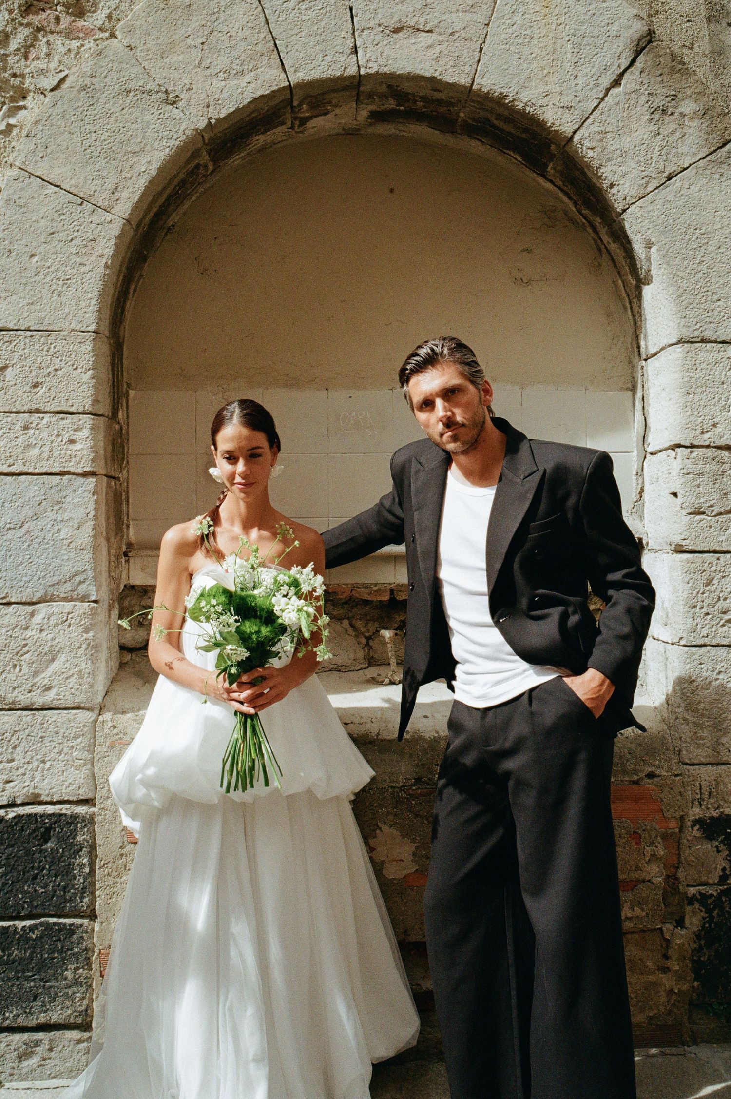 Bride holding green bouquet and groom in black suit pose inside a rustic stone archway together.