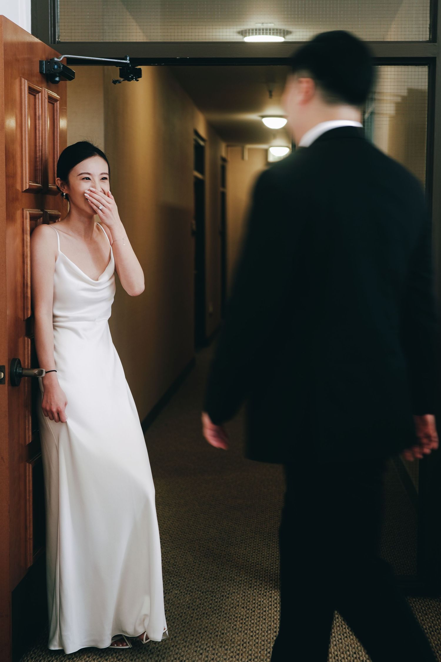 Emotional bride in white slip dress covers mouth as groom approaches in hotel corridor during first look moment.