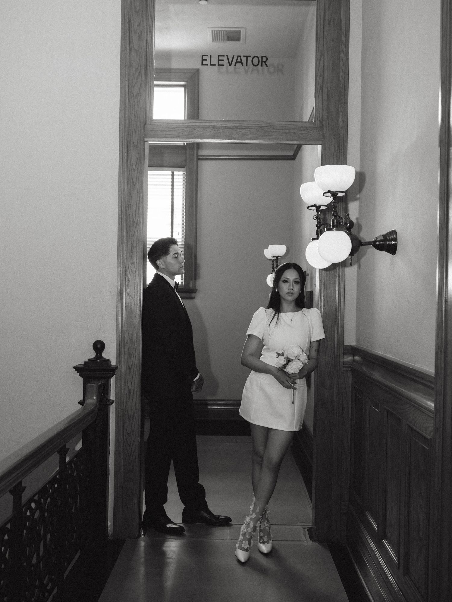 Black and white photo of bride and groom posing near an elevator sign in a classic building.