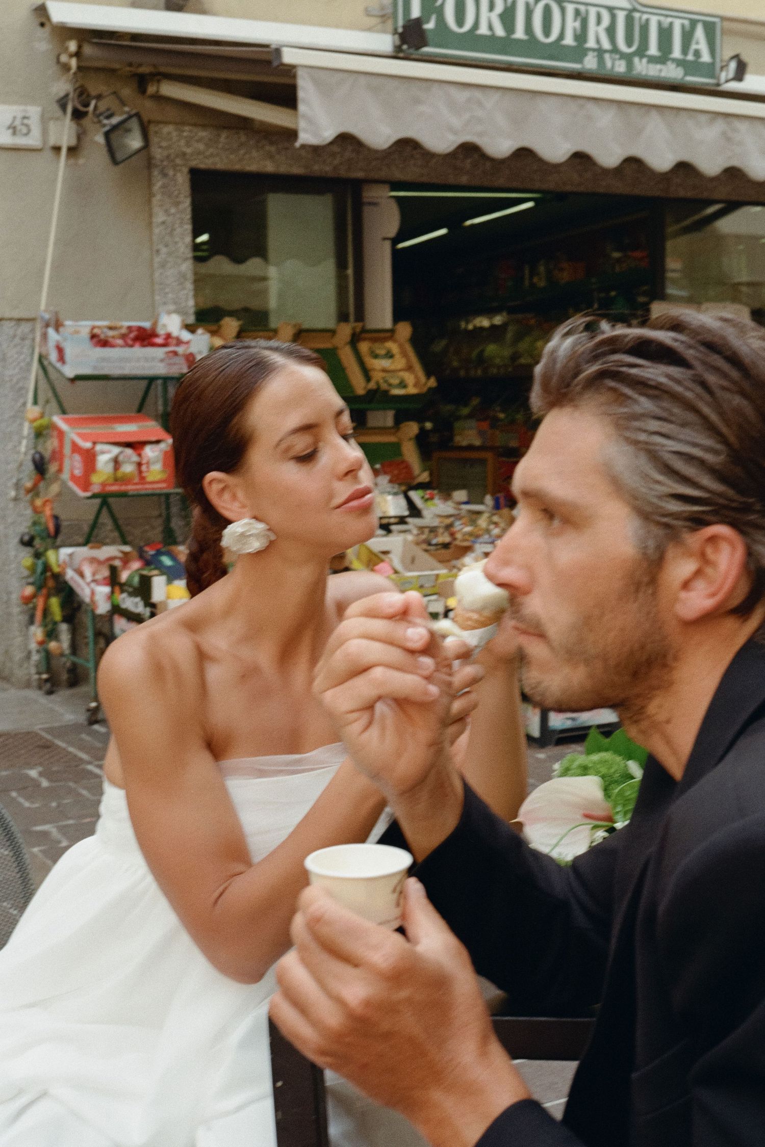 Bride and groom sharing an ice cream cone outside an Italian ortofrutta shop on their wedding day.