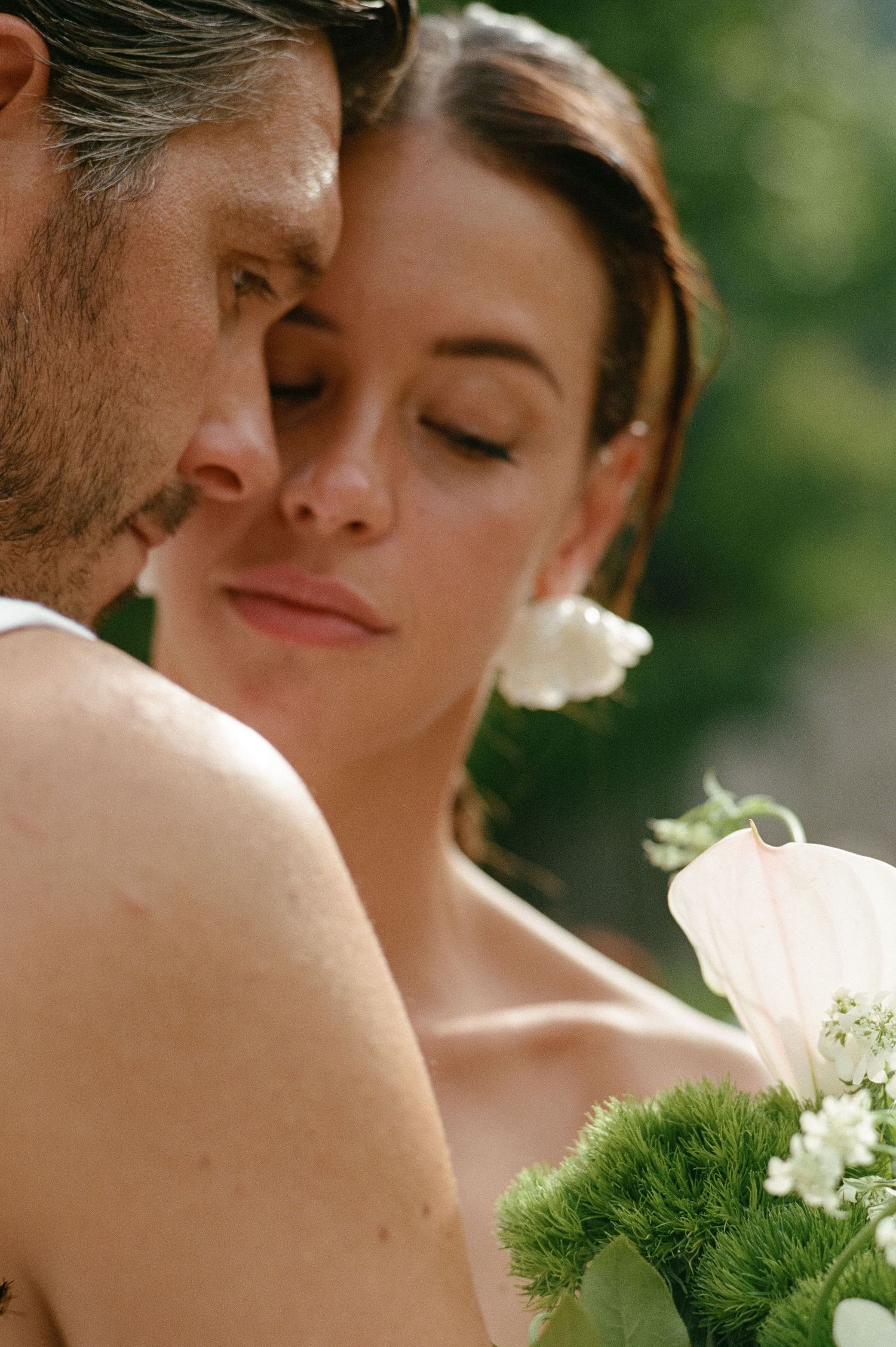 Bride with white flower in hair leans against groom holding elegant calla lily bouquet outdoors.