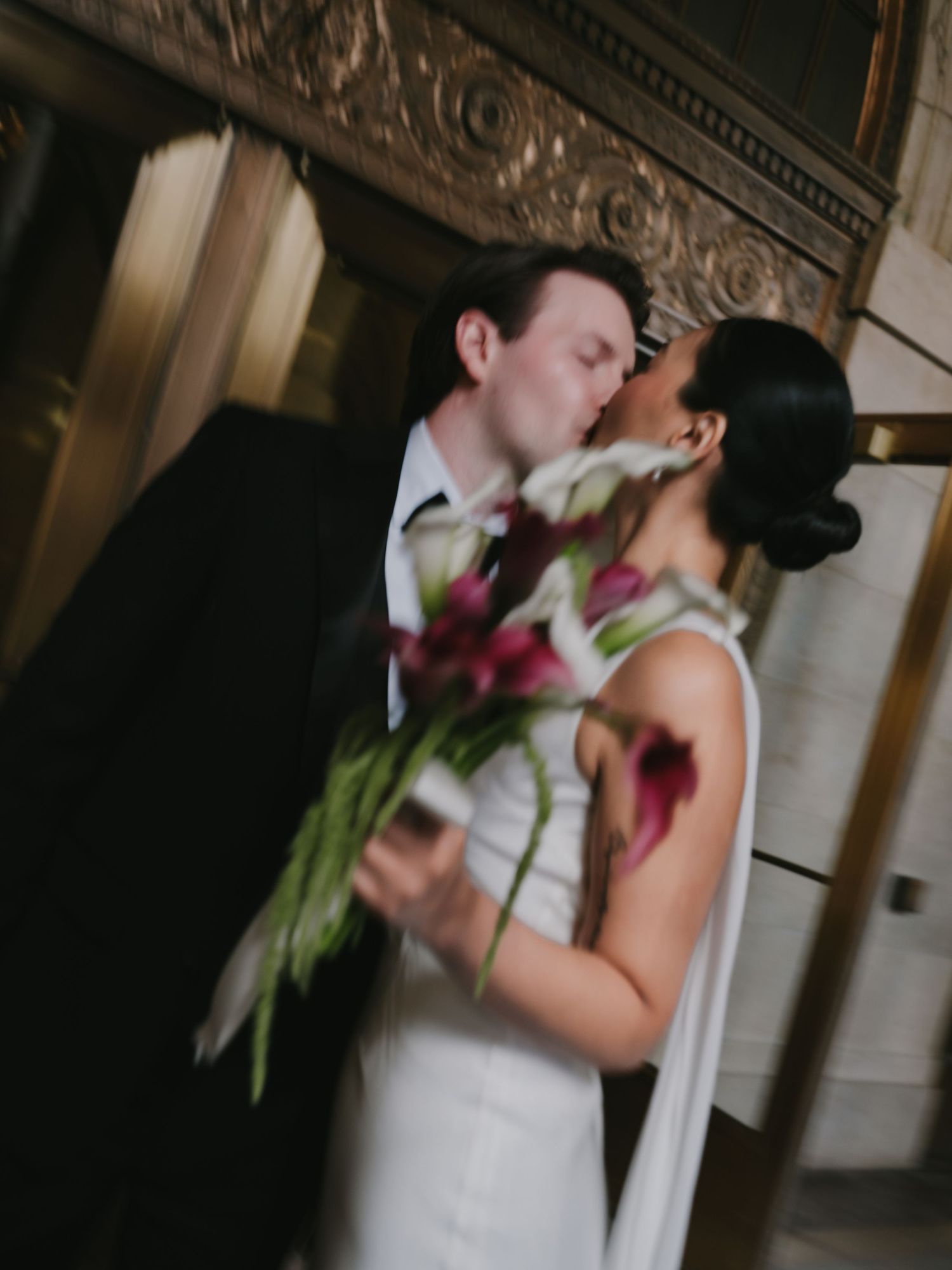 Blurred artistic shot of bride and groom sharing romantic first kiss holding calla lily bouquet.
