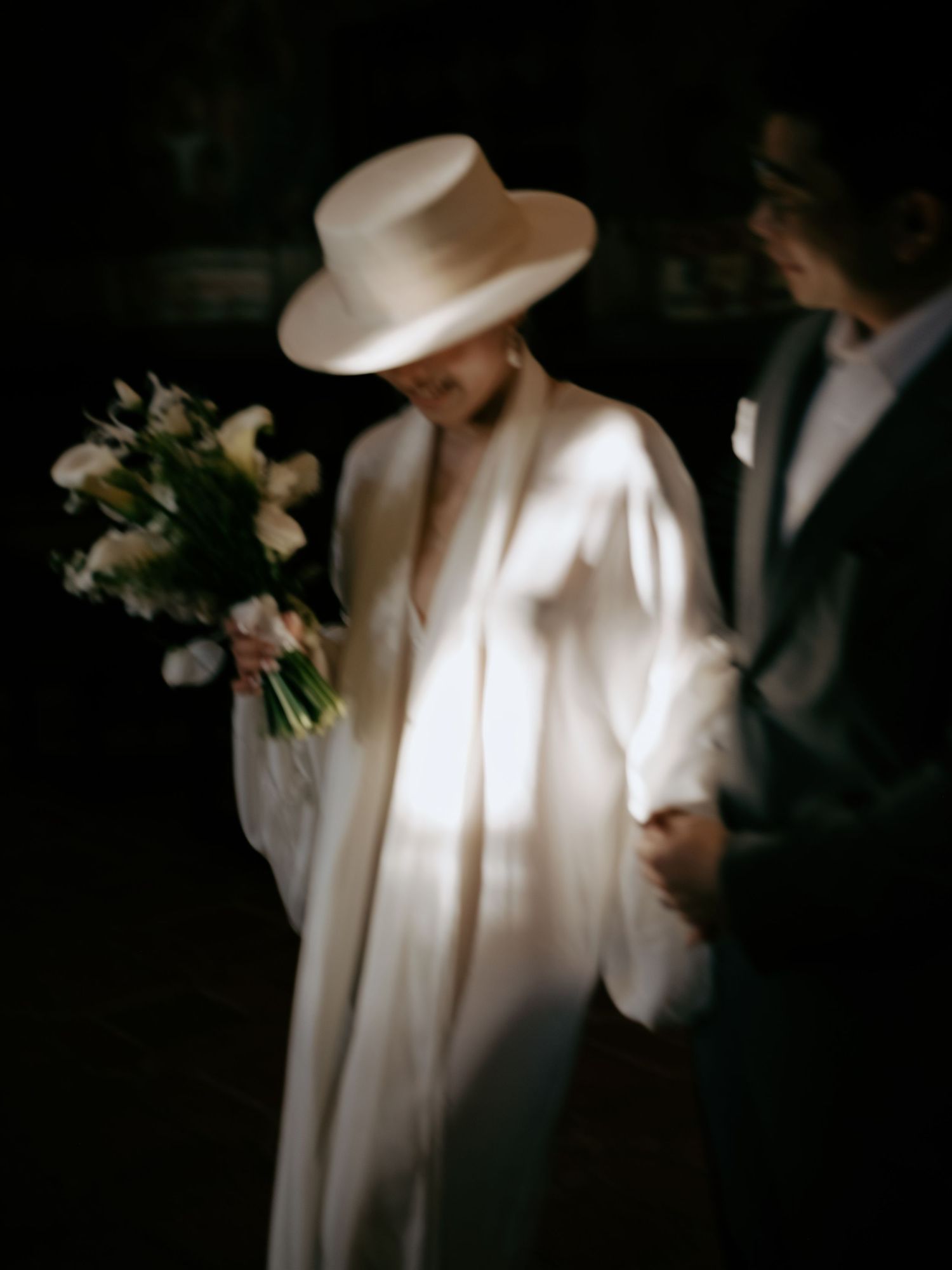 Artistic blurred photo of bride in white hat and gown holding bouquet beside suited groom.