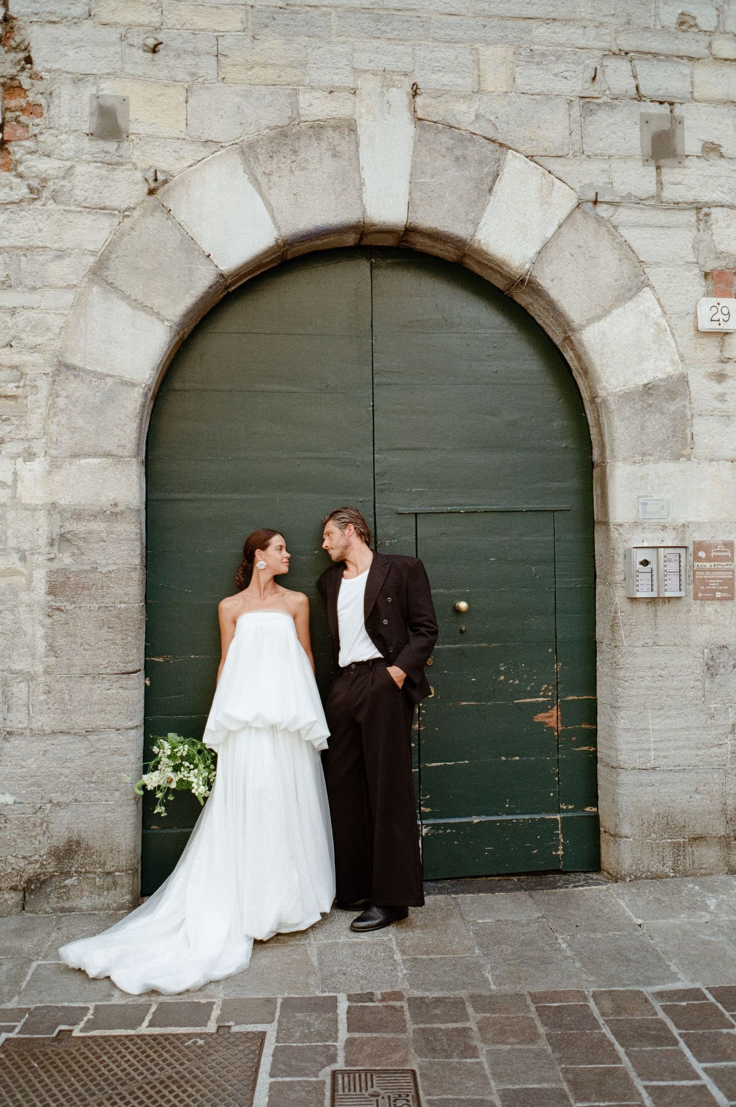 Bride in strapless white gown and groom in black suit standing by green arched wooden door.
