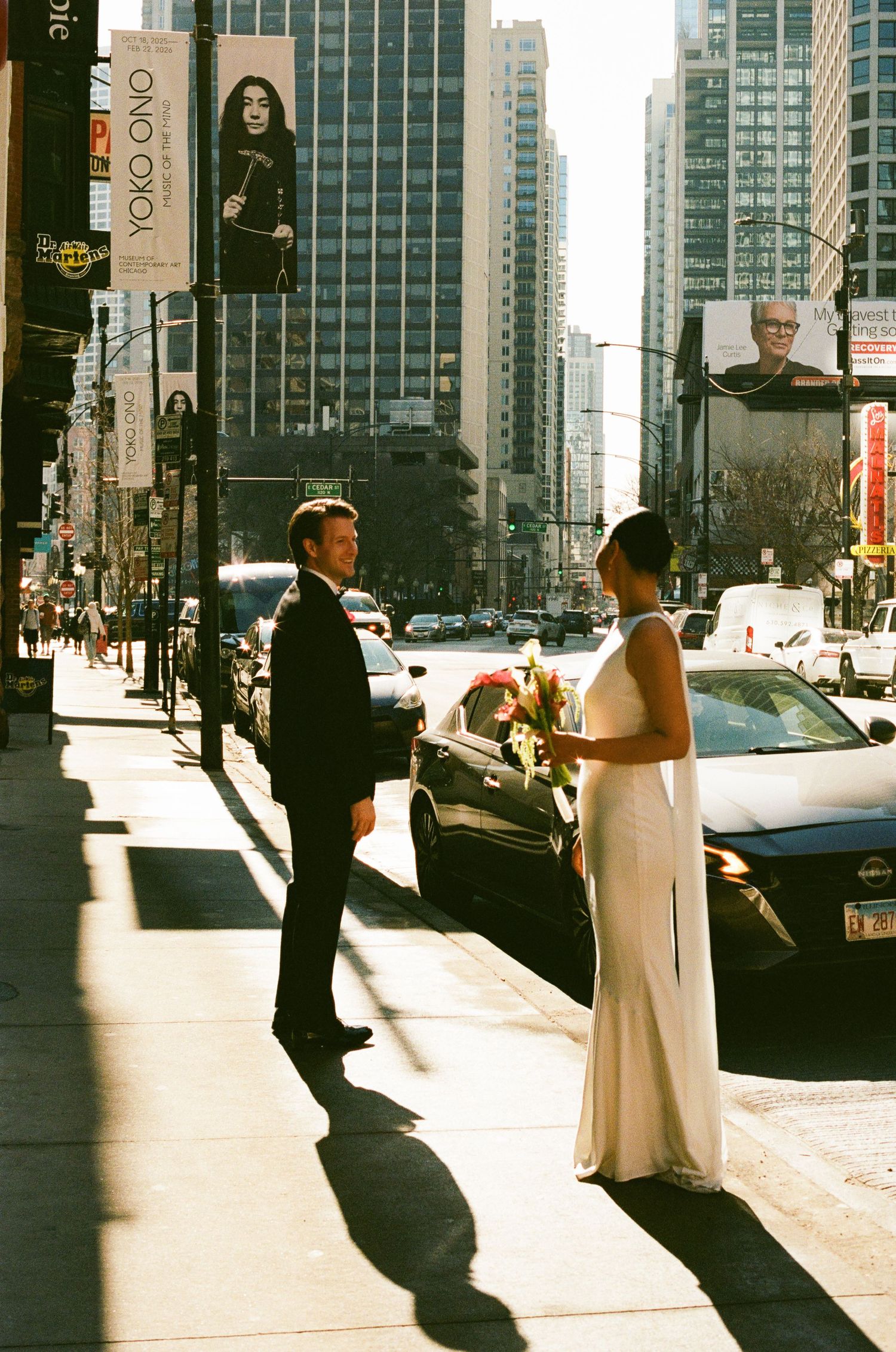Bride in sleek white gown and groom in tuxedo face each other on sunny Chicago city street.