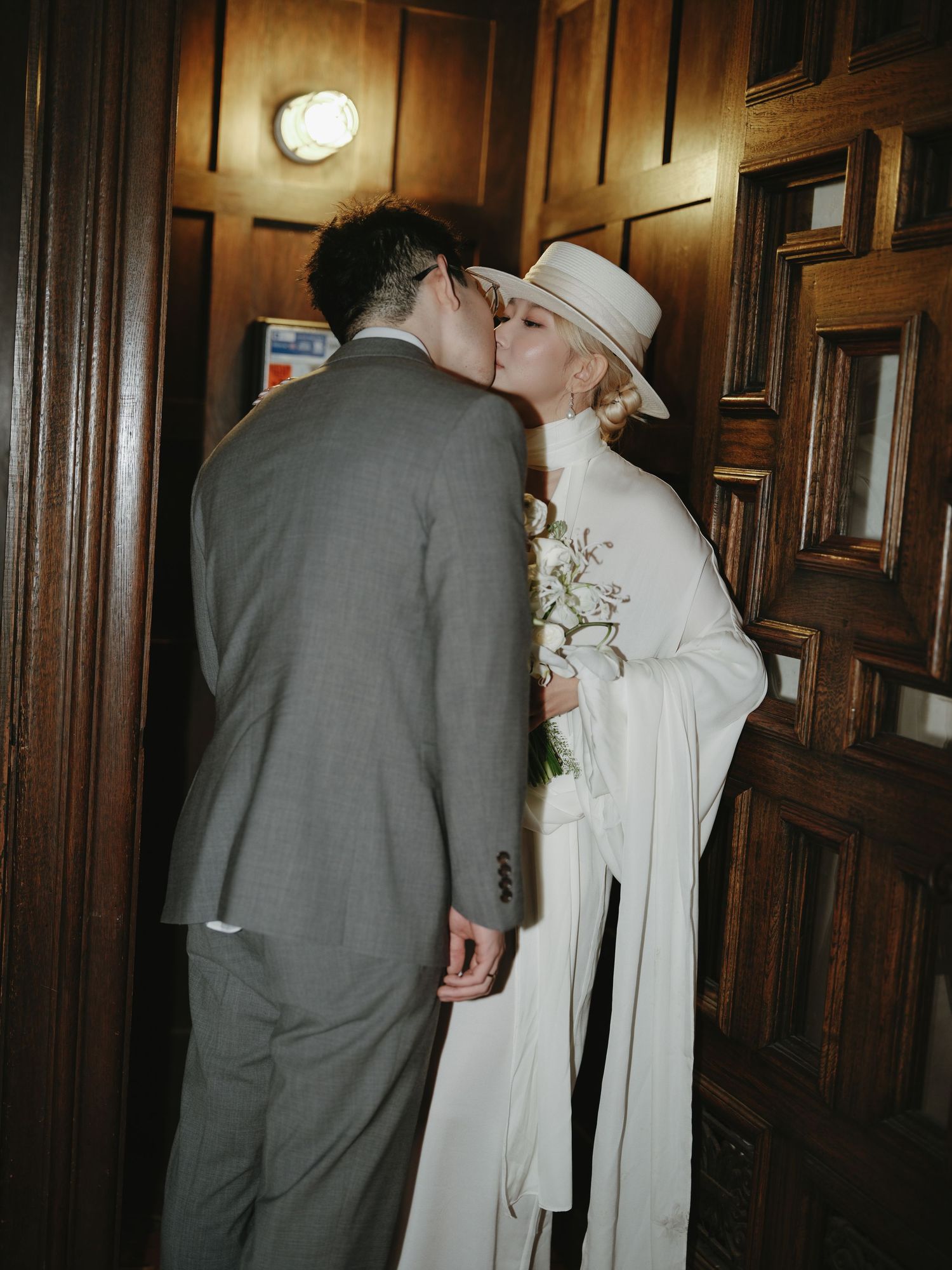 Bride in white wide-brim hat and groom in gray suit share a kiss by wooden doors.
