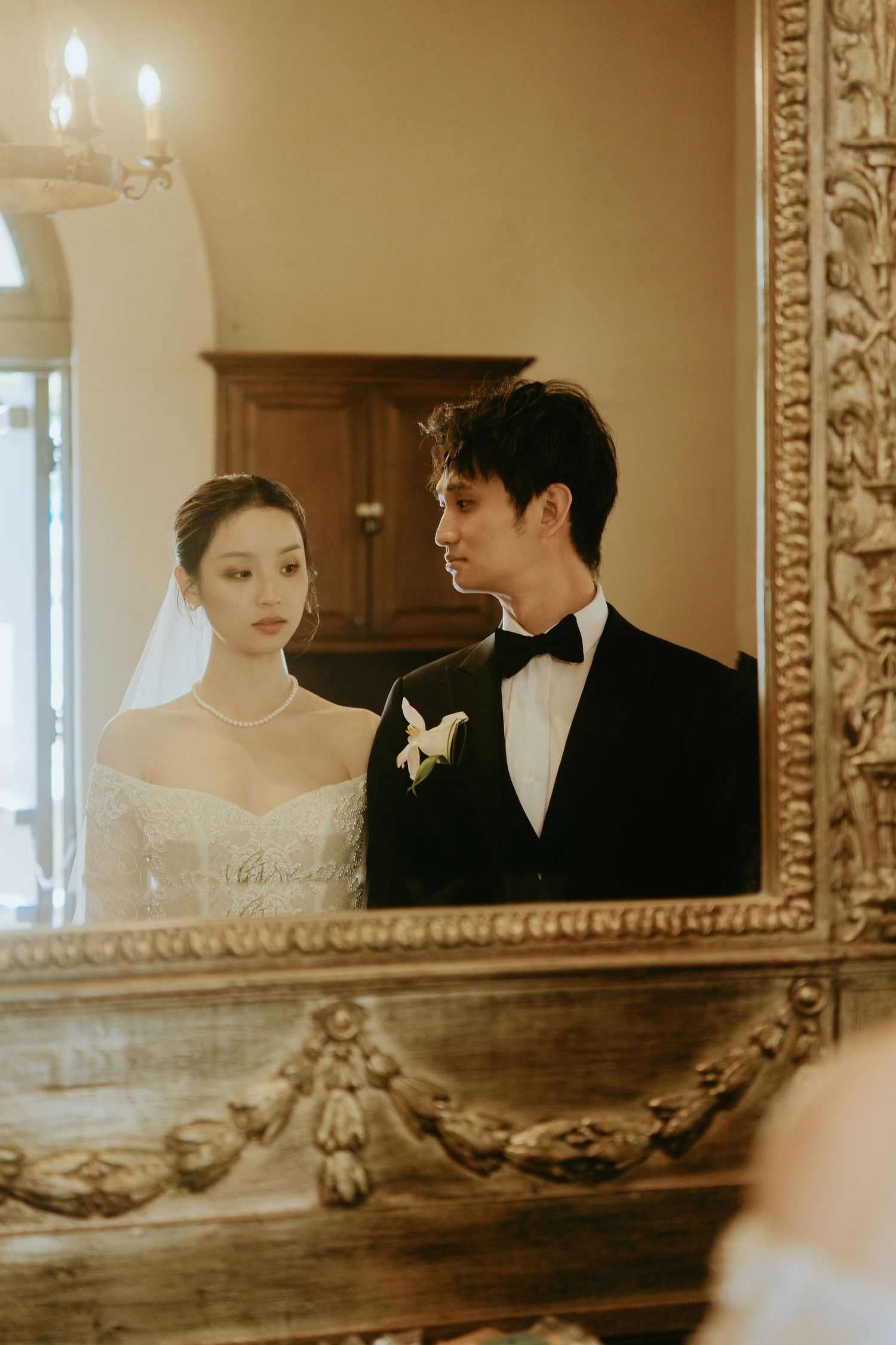 Bride and groom in elegant wedding attire reflected in ornate gold-framed vintage mirror.
