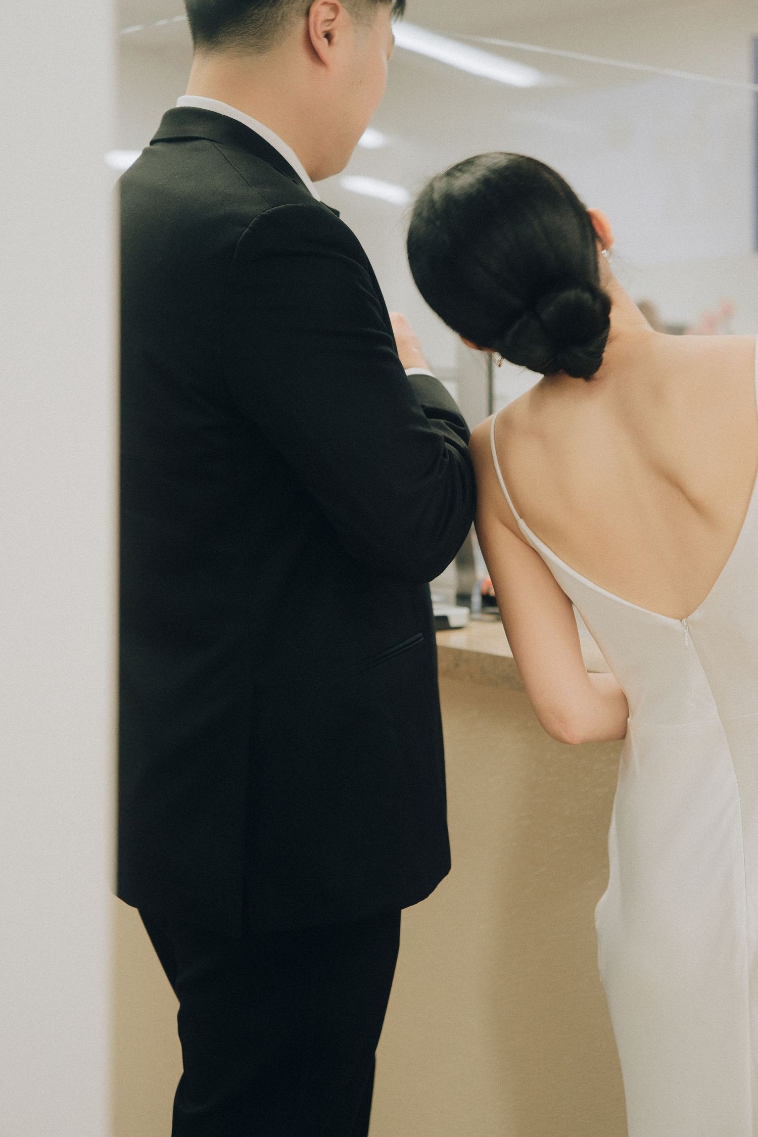 Groom in black suit helping bride in white backless dress at mirror.