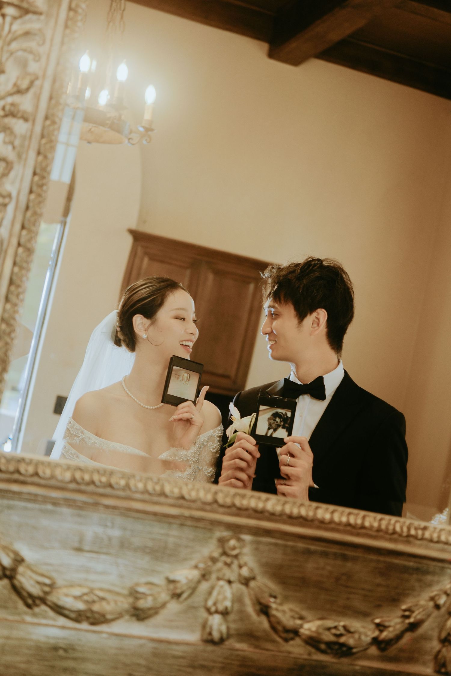 Smiling bride and groom holding Polaroid photos reflected in ornate gold mirror at wedding.