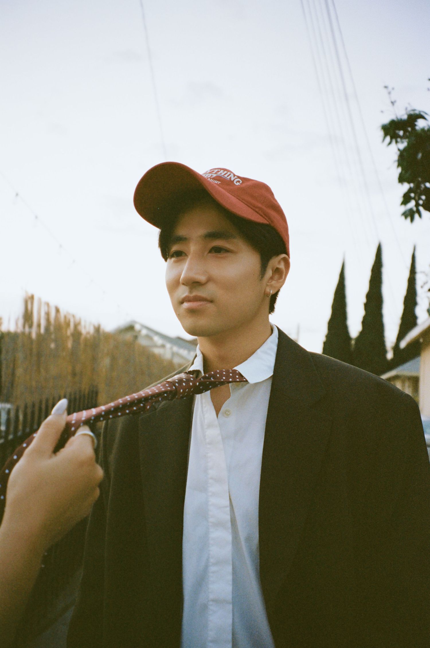 Young Asian man in red cap and black blazer with polka dot tie outdoors.