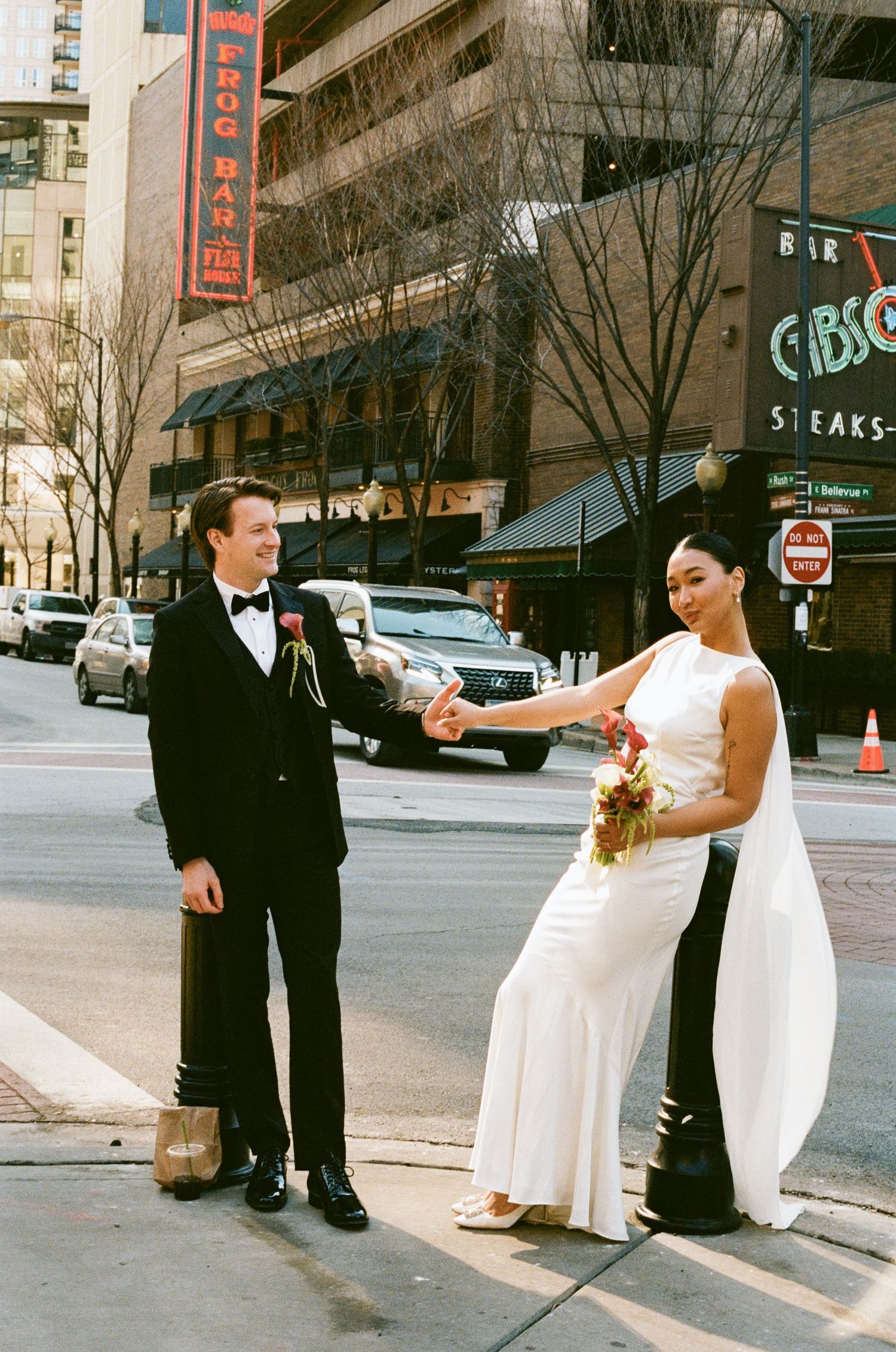 Bride in white gown and groom in black tuxedo hold hands on a city street in Chicago.