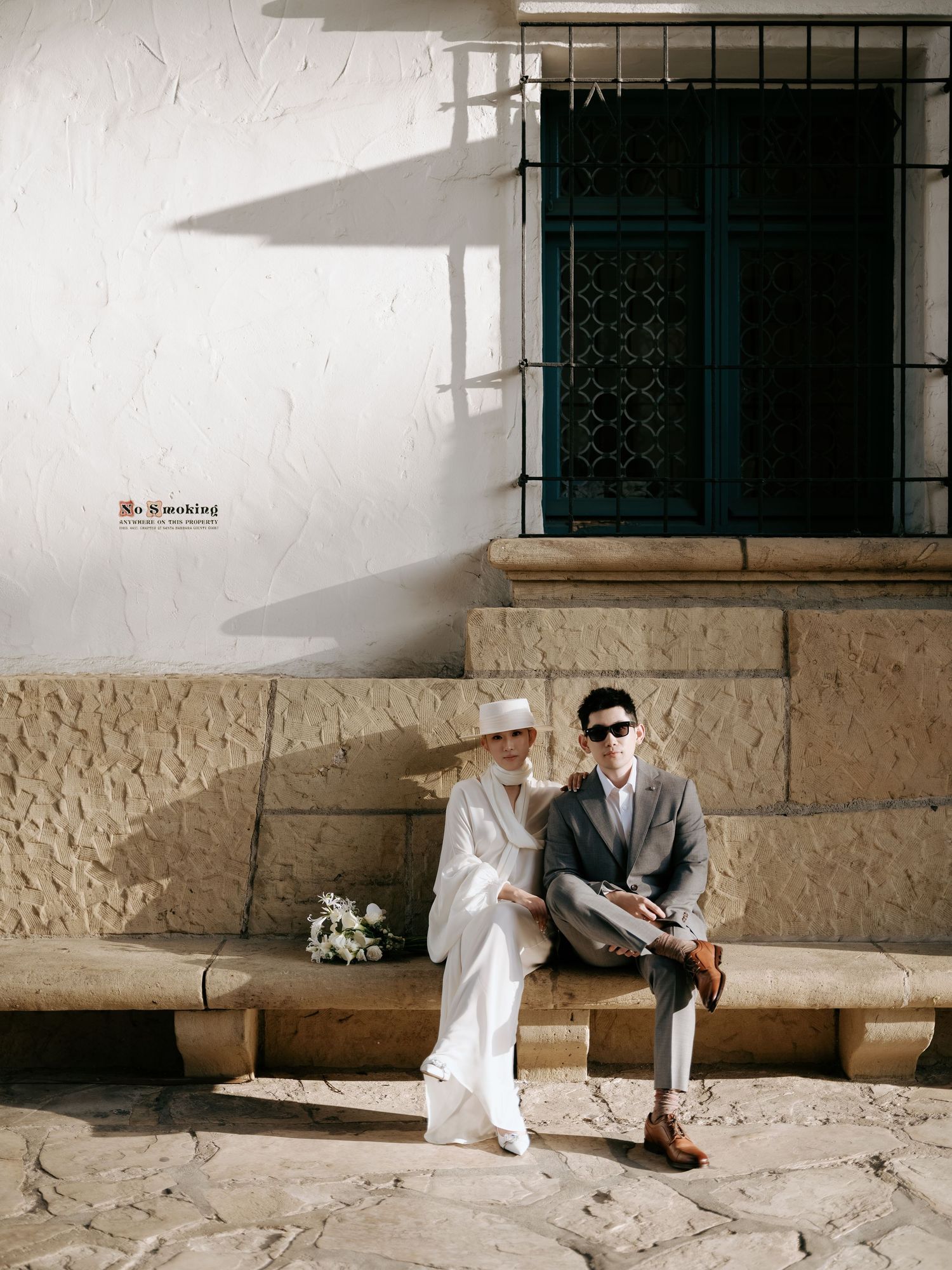 Stylish bride in white wide-brim hat and silk outfit sits with groom on stone bench in sunny Mediterranean setting.