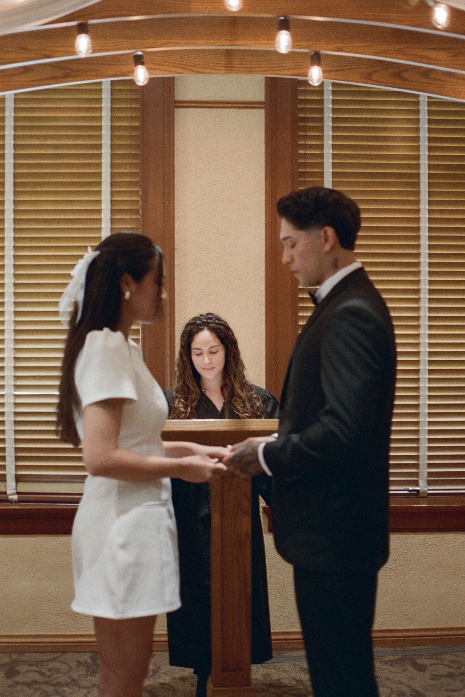 Bride in white mini dress and groom in black tuxedo hold hands during wedding ceremony.