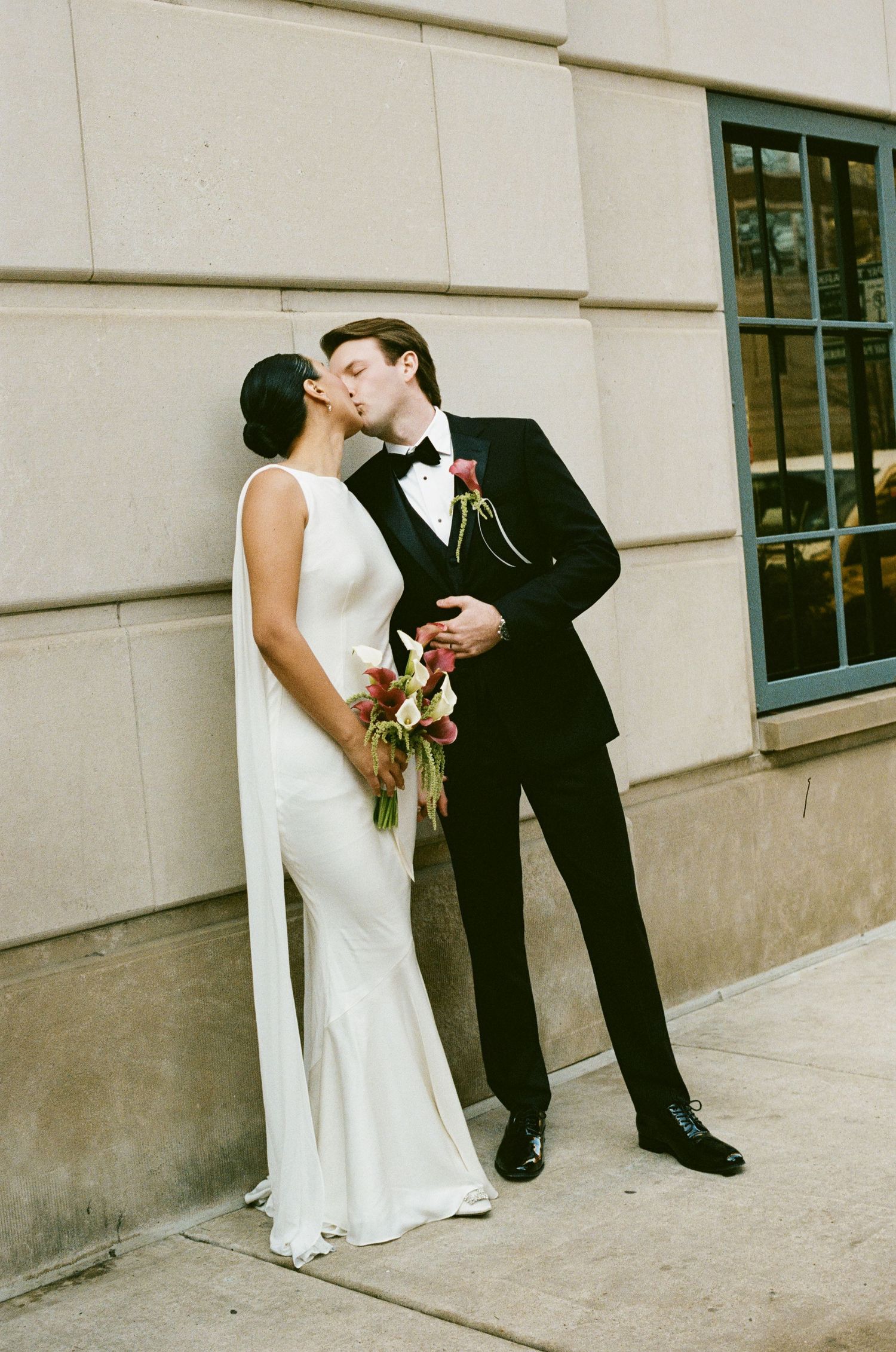 Bride and groom share a kiss against stone building wall, bride holding colorful wedding bouquet.