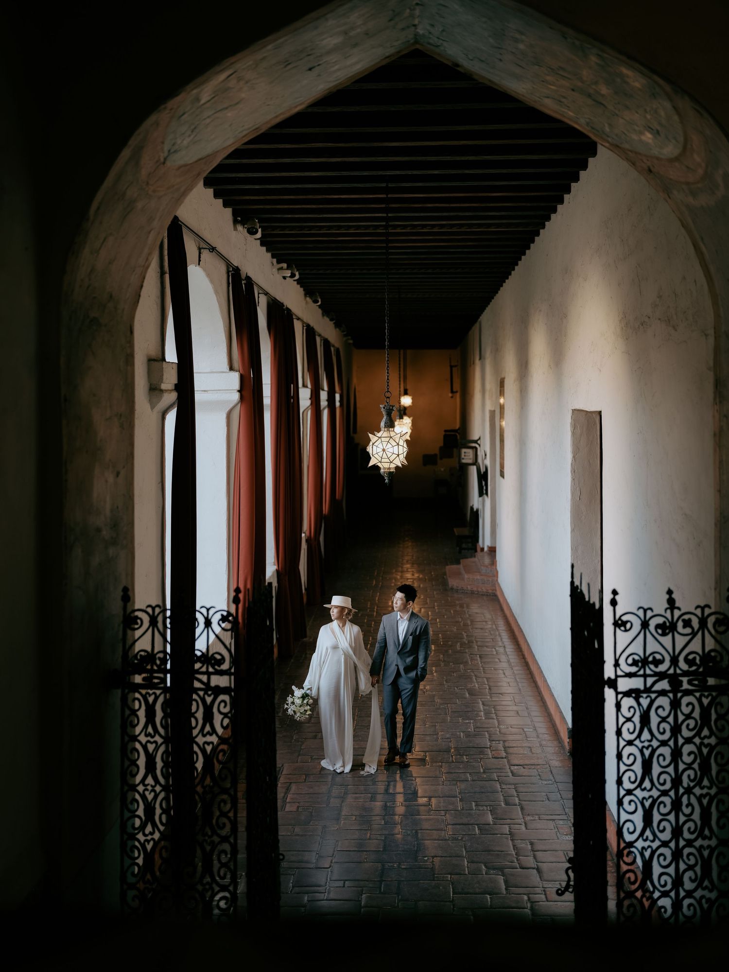 Wedding couple walking through ornate iron gate in terracotta-tiled corridor with hanging lantern.