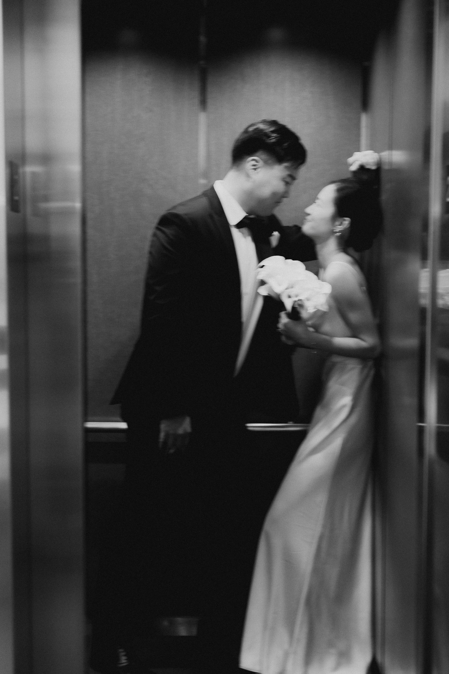 Black and white romantic photo of Asian bride and groom sharing intimate moment in elevator on wedding day.