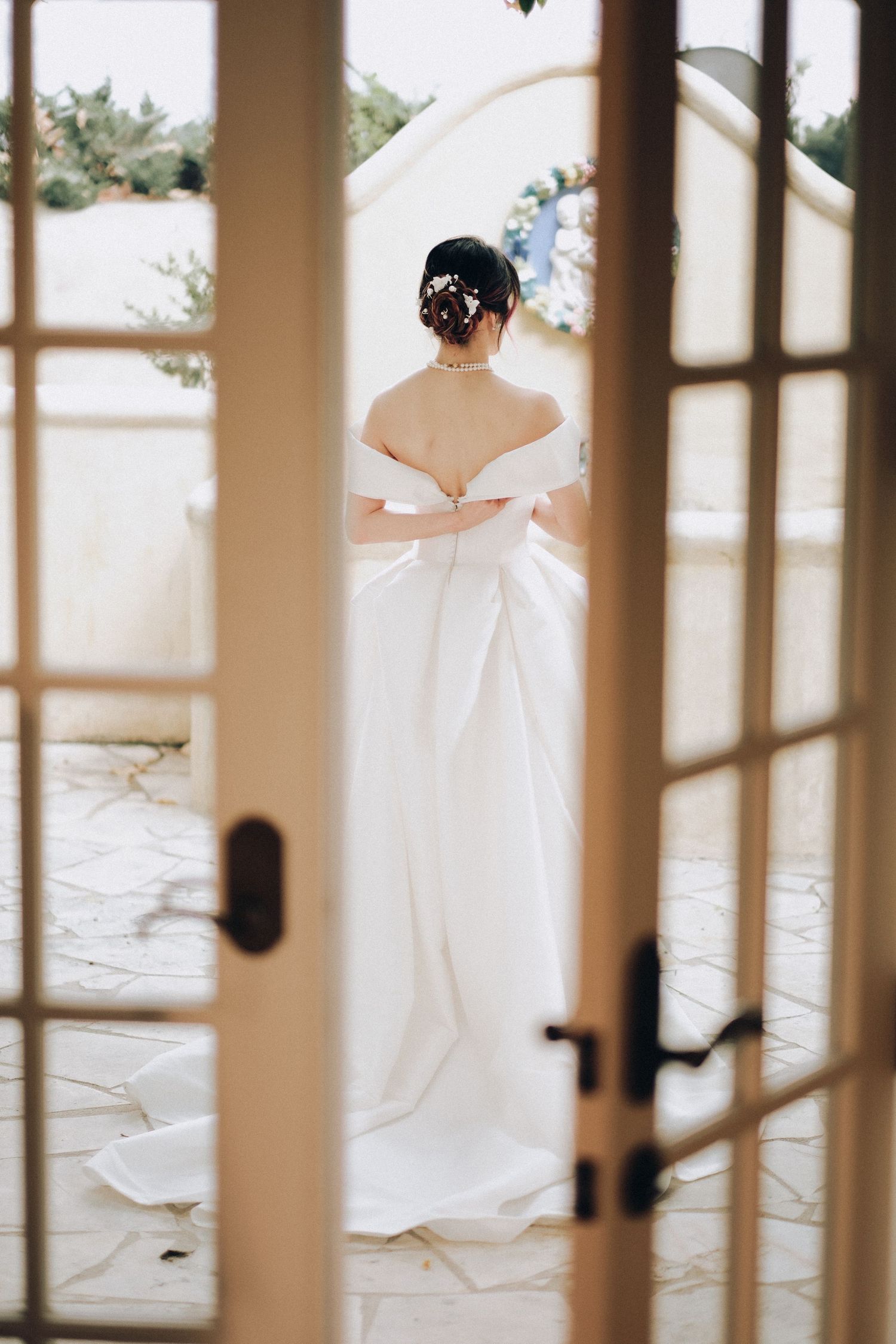 Bride in off-shoulder white wedding gown viewed from behind through French doors.