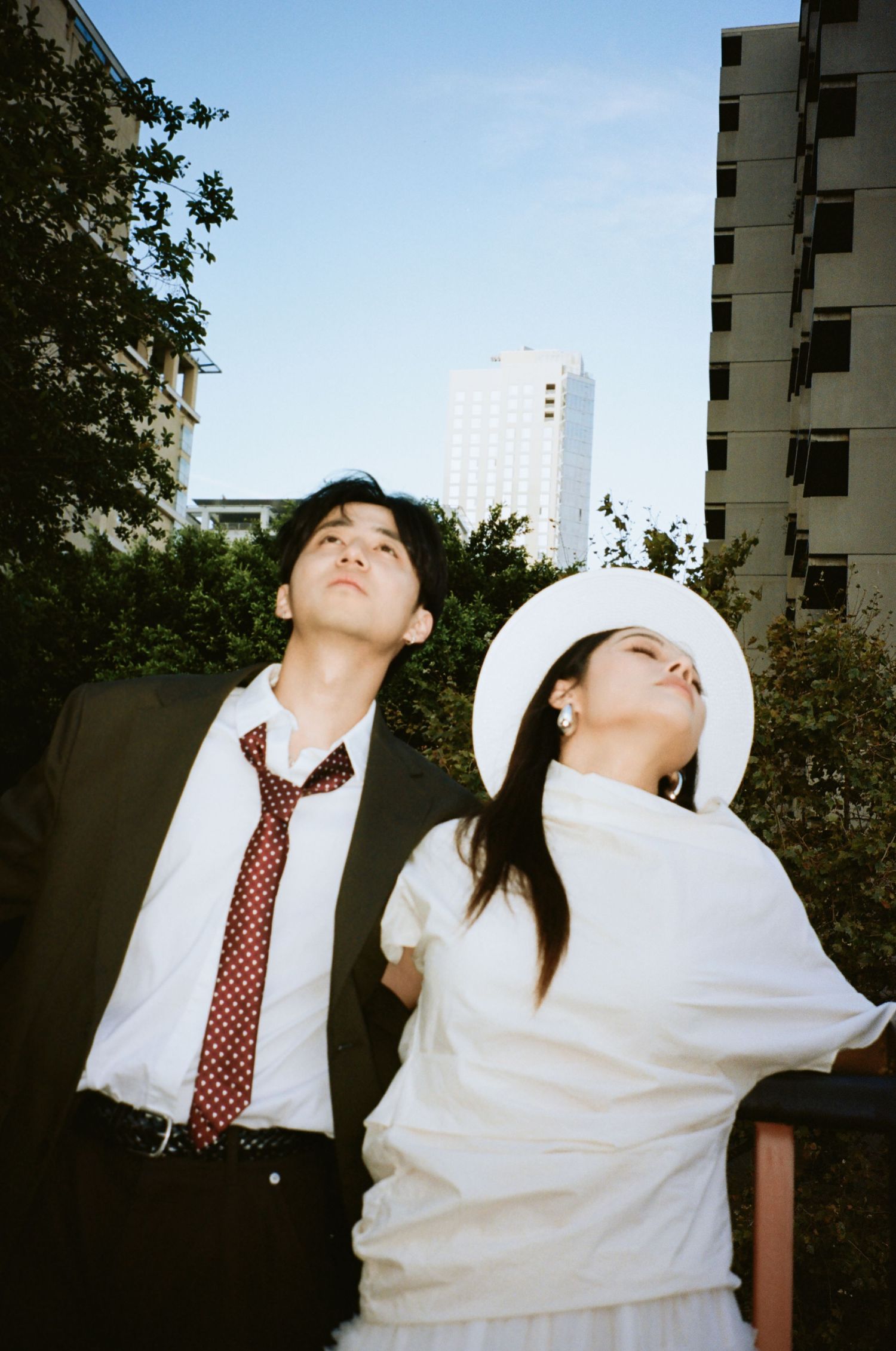 Wedding couple in white and dark suits lean back dramatically against urban building backdrop in bright daylight.