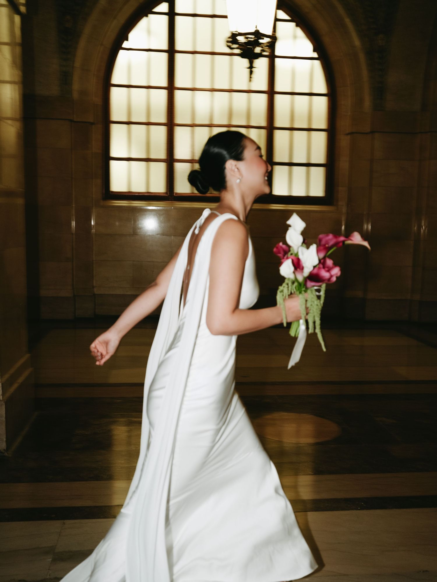 Elegant bride in sleek white gown with cape back walks through grand hall holding pink calla lily bouquet.