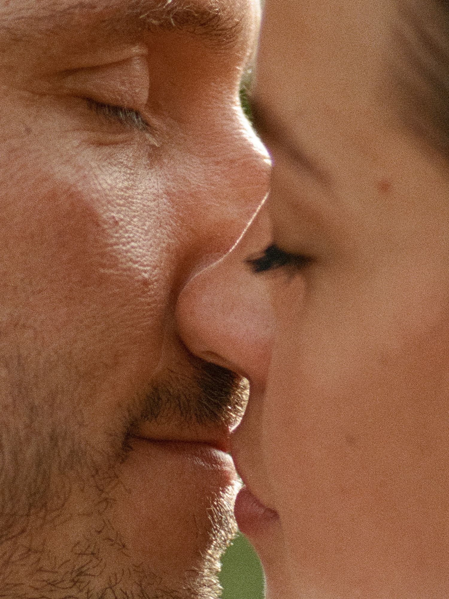 Extreme close-up profile of a couple sharing a gentle kiss in warm natural sunlight.