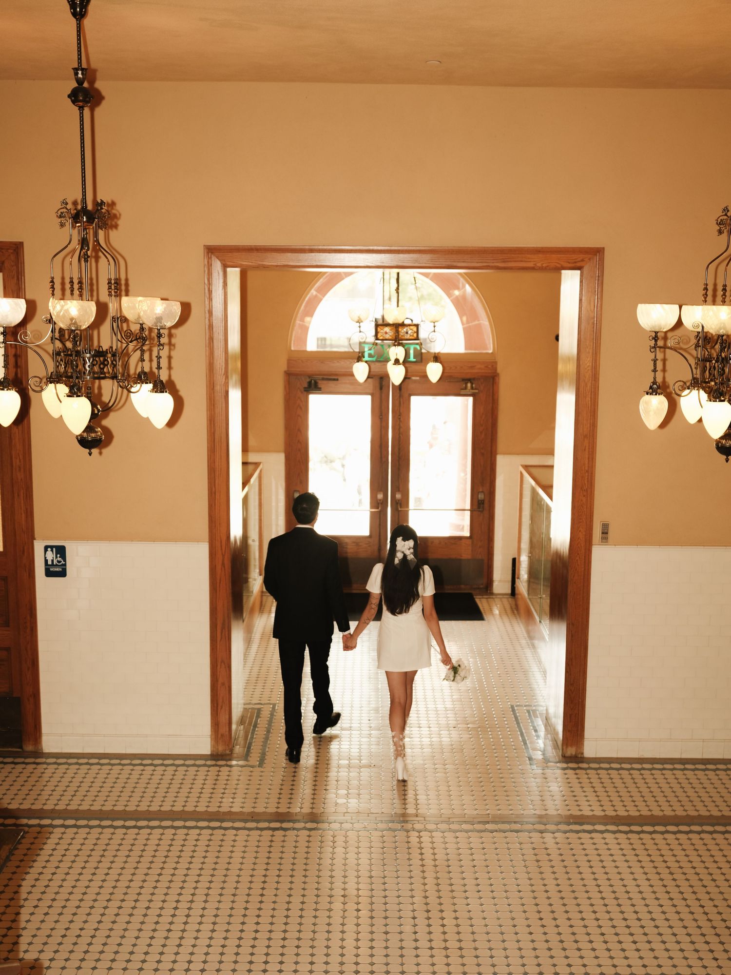 Bride and groom walk hand-in-hand through elegant hallway with chandeliers toward arched doors.