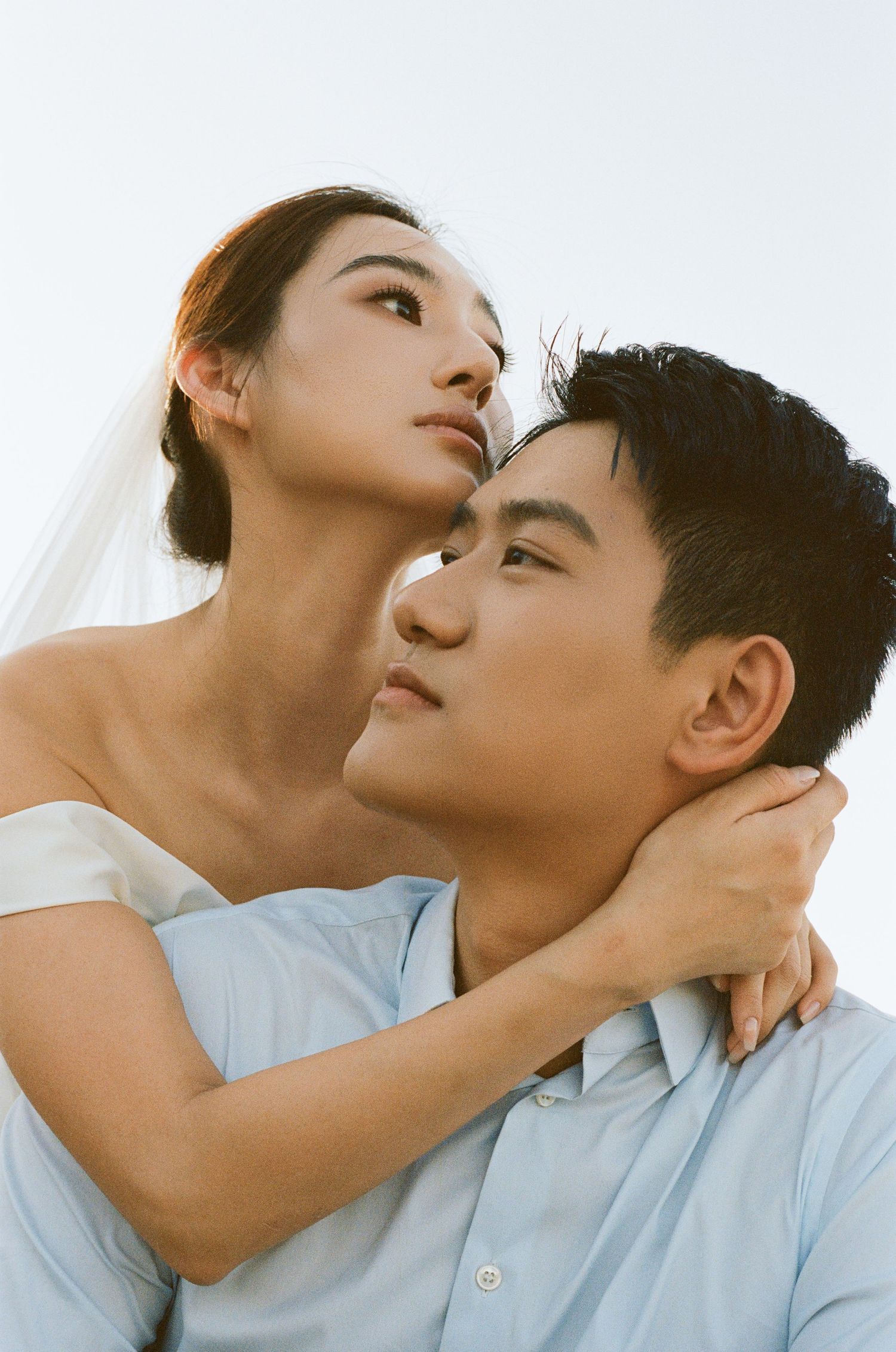 Bride in off-shoulder gown embraces groom from behind as both gaze upward against a pale sky.
