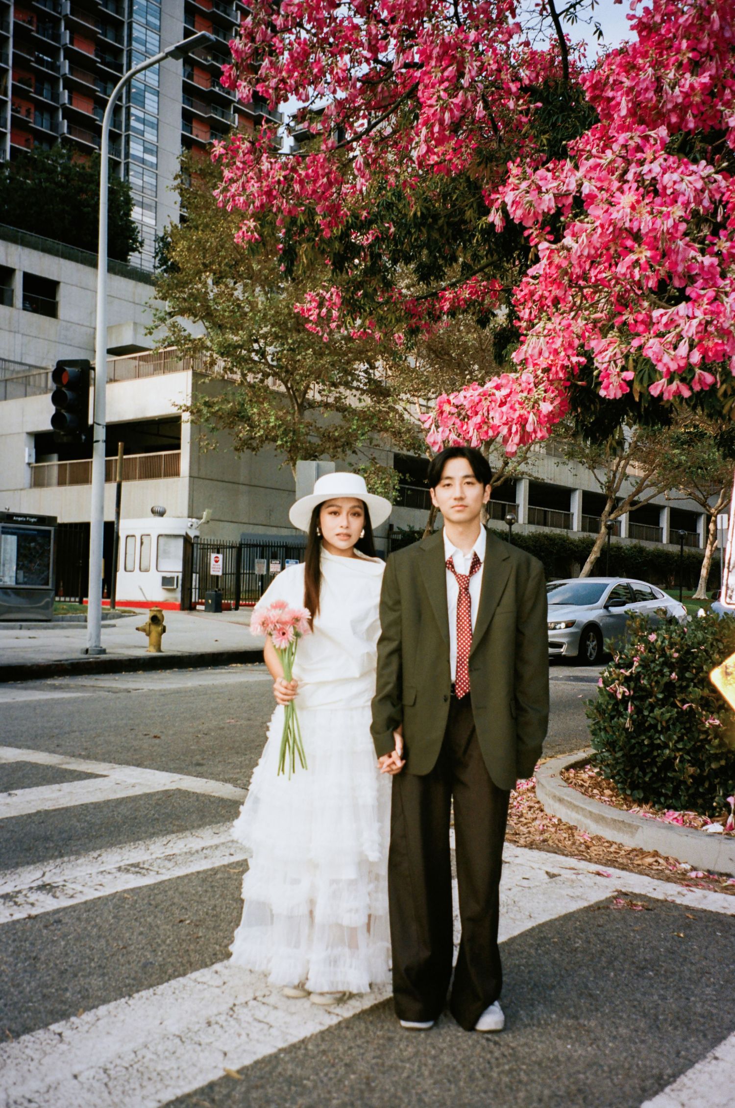 Couple holds hands on crosswalk beneath blooming pink trees, woman in white dress with hat, man in olive suit.