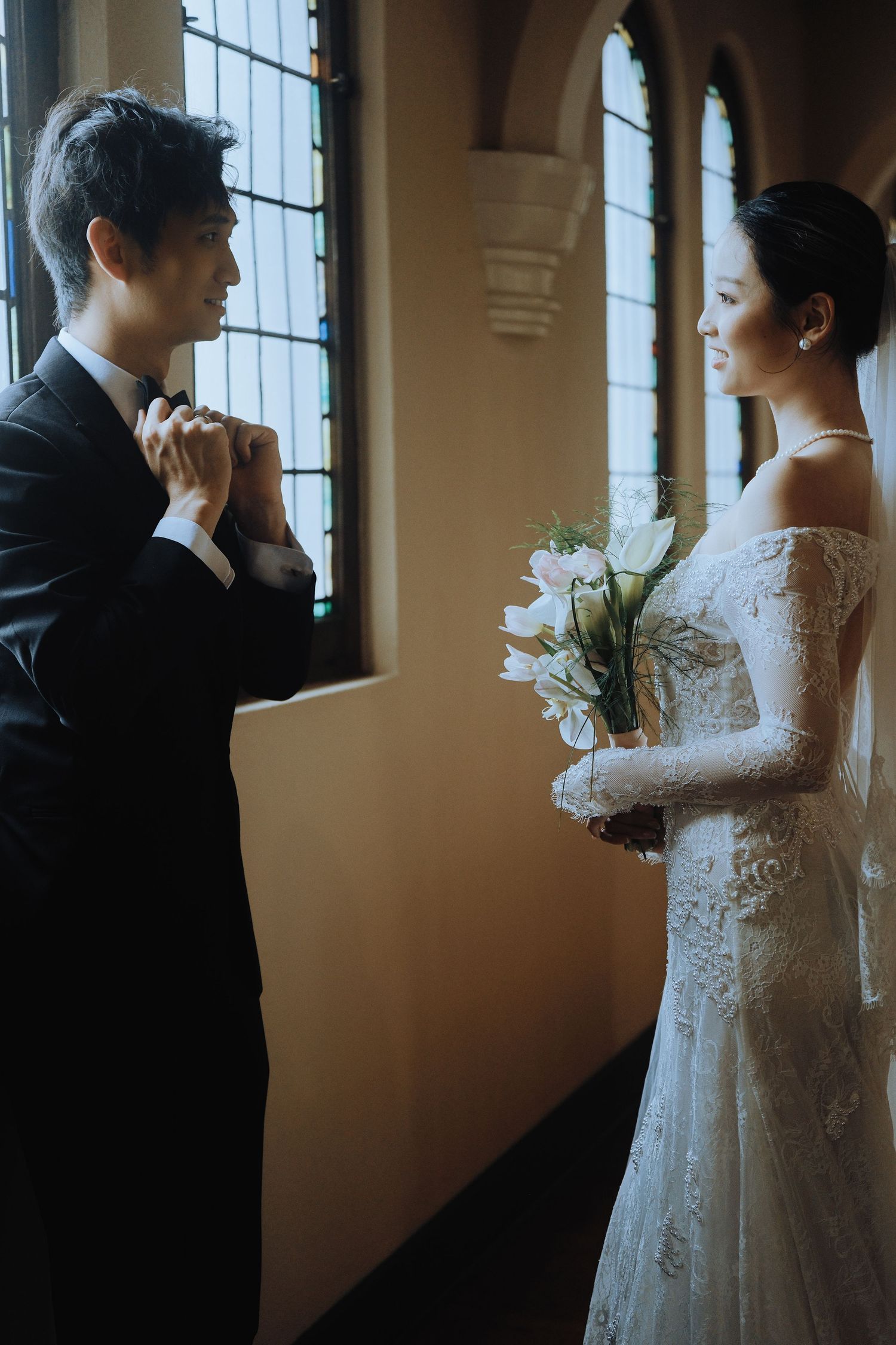 Groom clasps brides hands tenderly as they face each other by stained glass church windows.