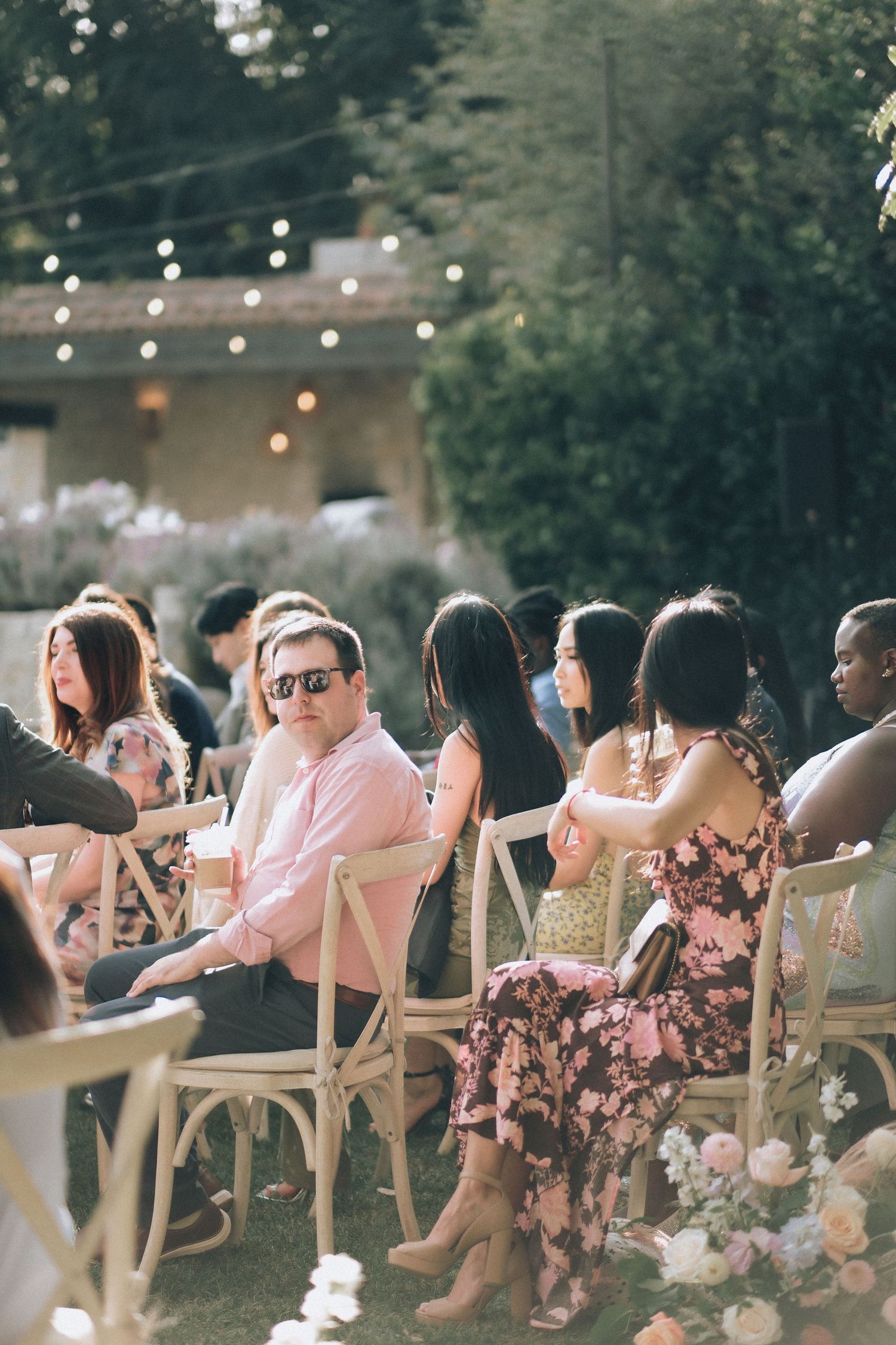 Wedding guests seated in white crossback chairs at an outdoor garden ceremony with warm string lights above.