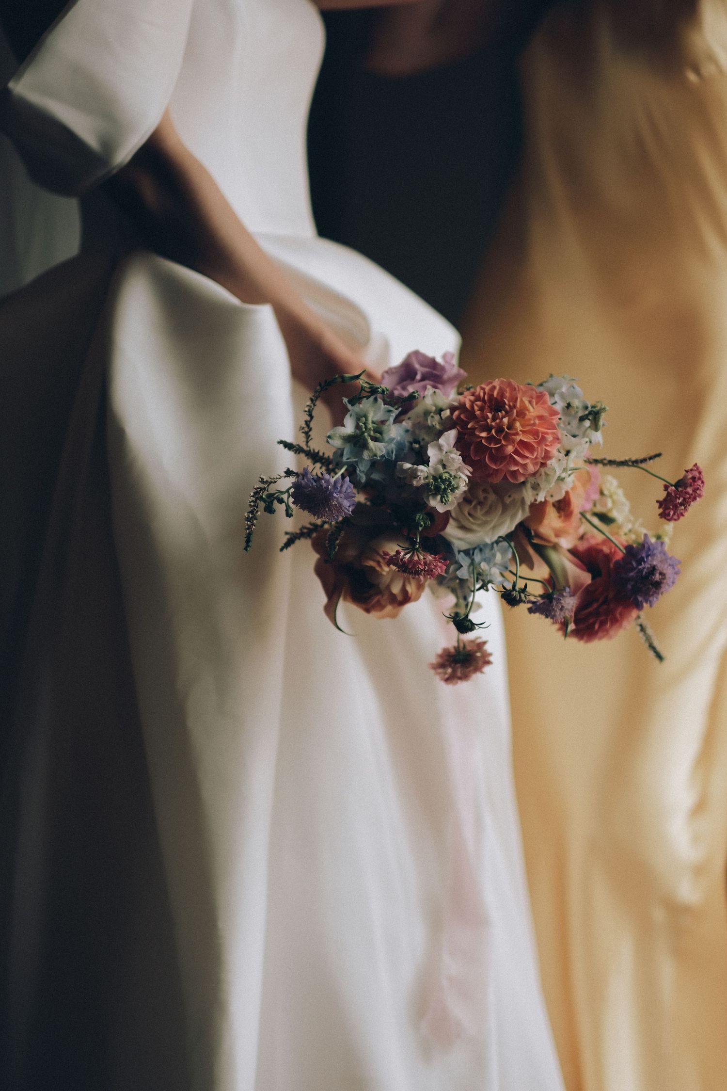 Two women in white and yellow gowns holding a colorful wedding bouquet with dahlias, purple flowers, and greenery.